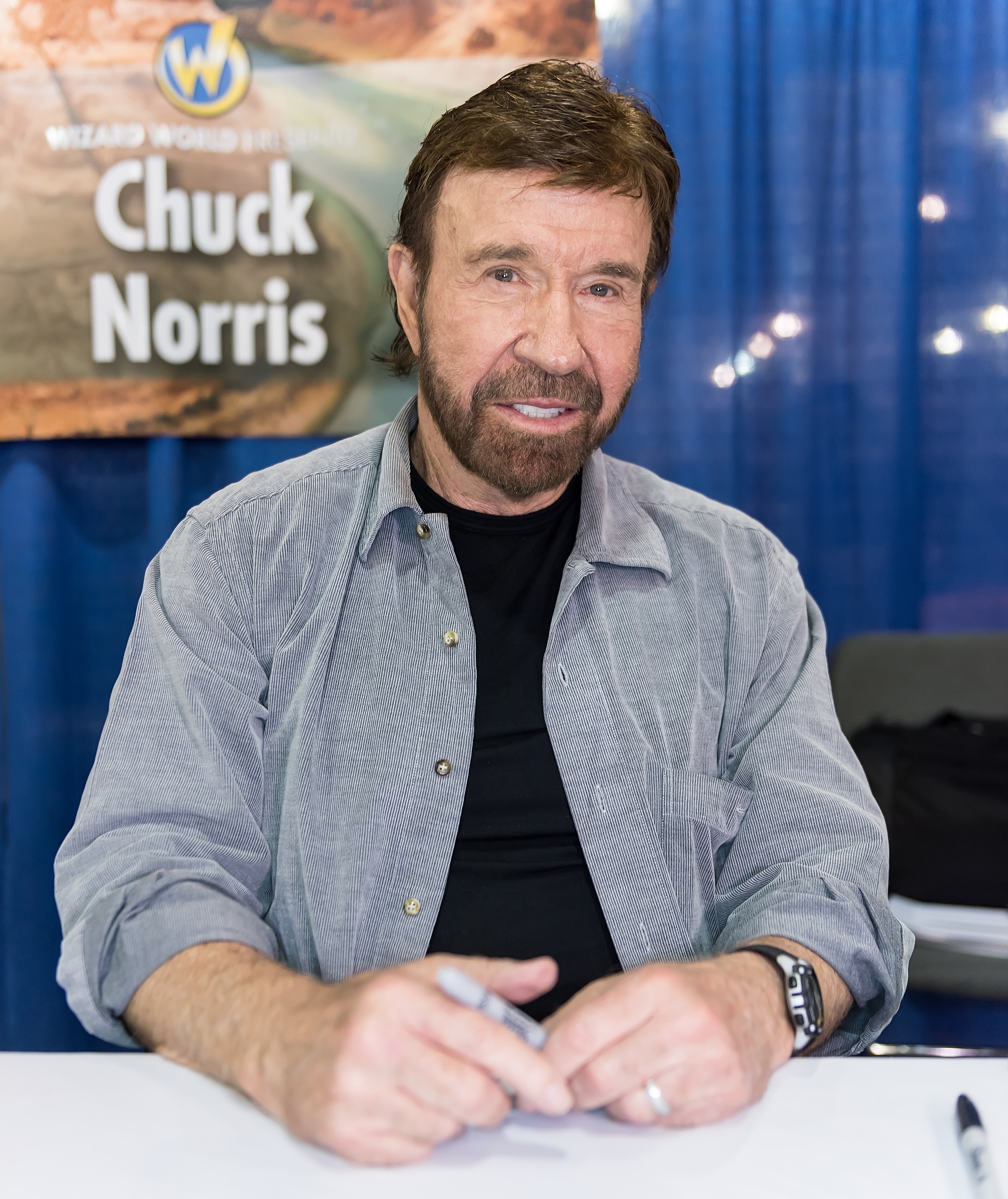 Man with a beard and casual garment  signing autographs astatine  a nationalist   event. Poster down  him has words "Wizard World Comic Con Chuck Norris."