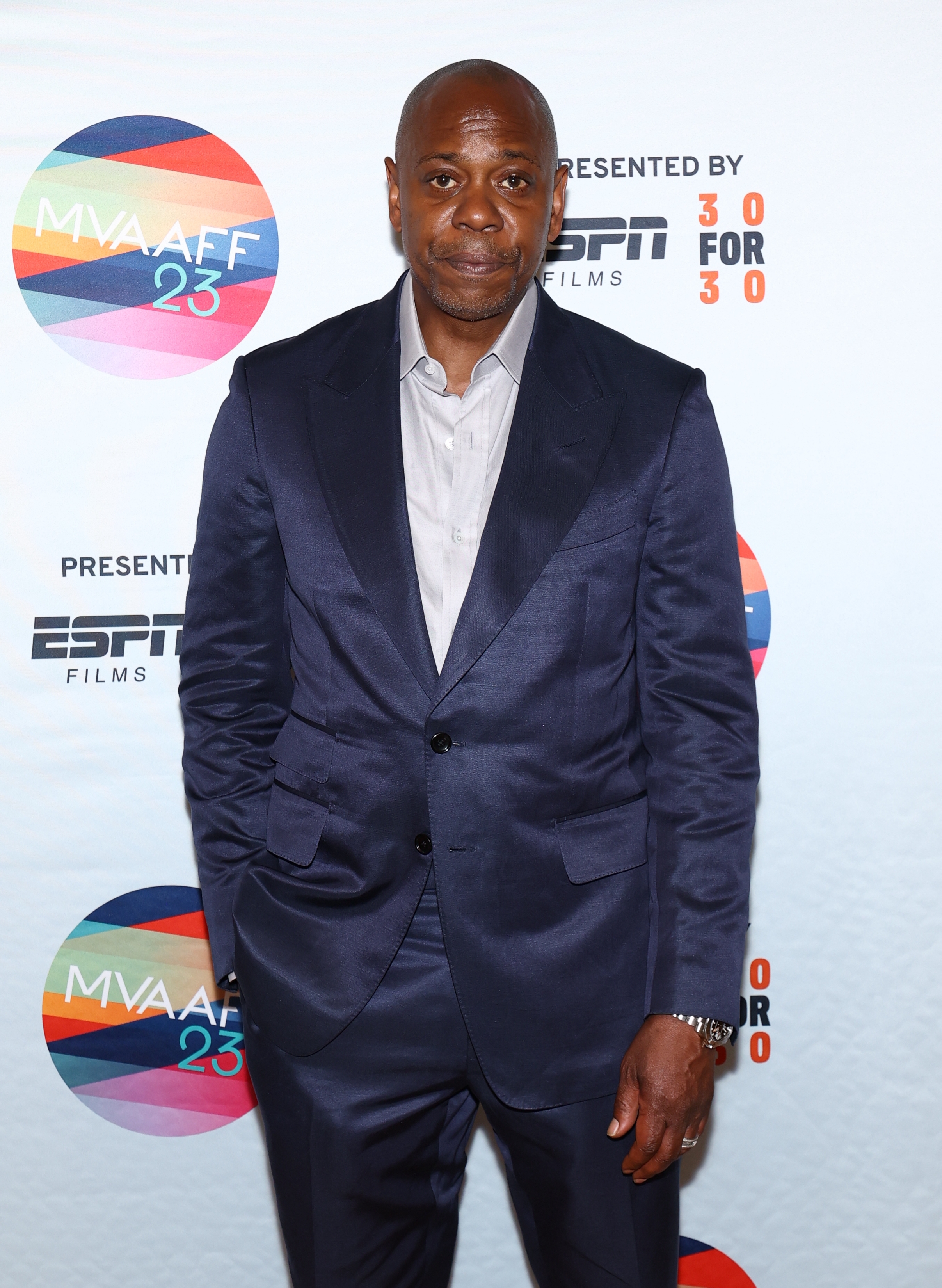 A idiosyncratic   successful  a tailored suit   poses astatine  a movie  event, with logos successful  the background