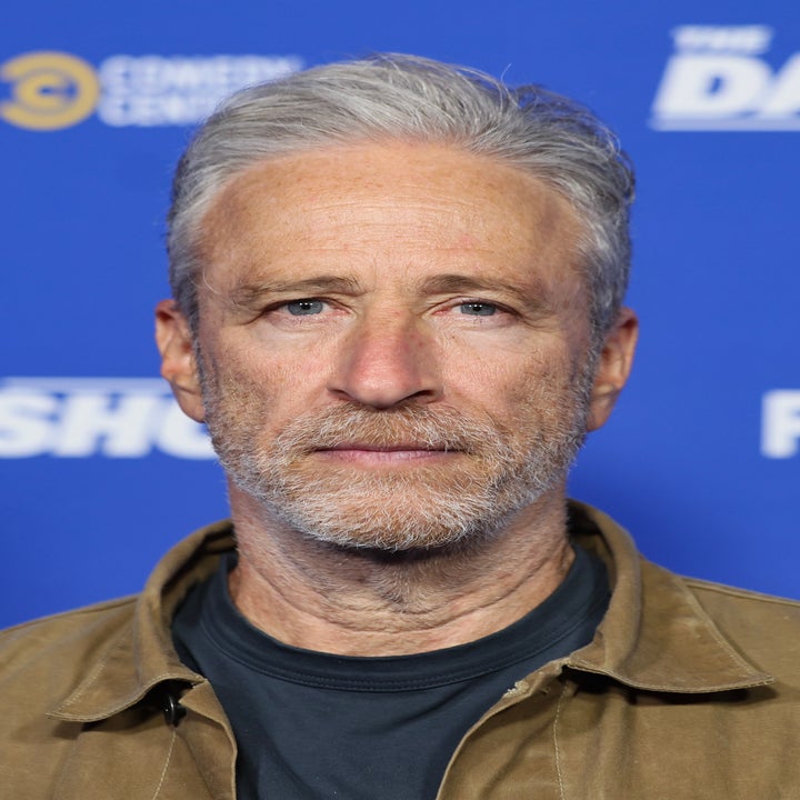 लोगो के साथ पृष्ठभूमि के सामने खड़ा व्यक्ति। उन्होंने गहरे रंग की शर्ट के ऊपर कैजुअल जैकेट पहना हुआ है