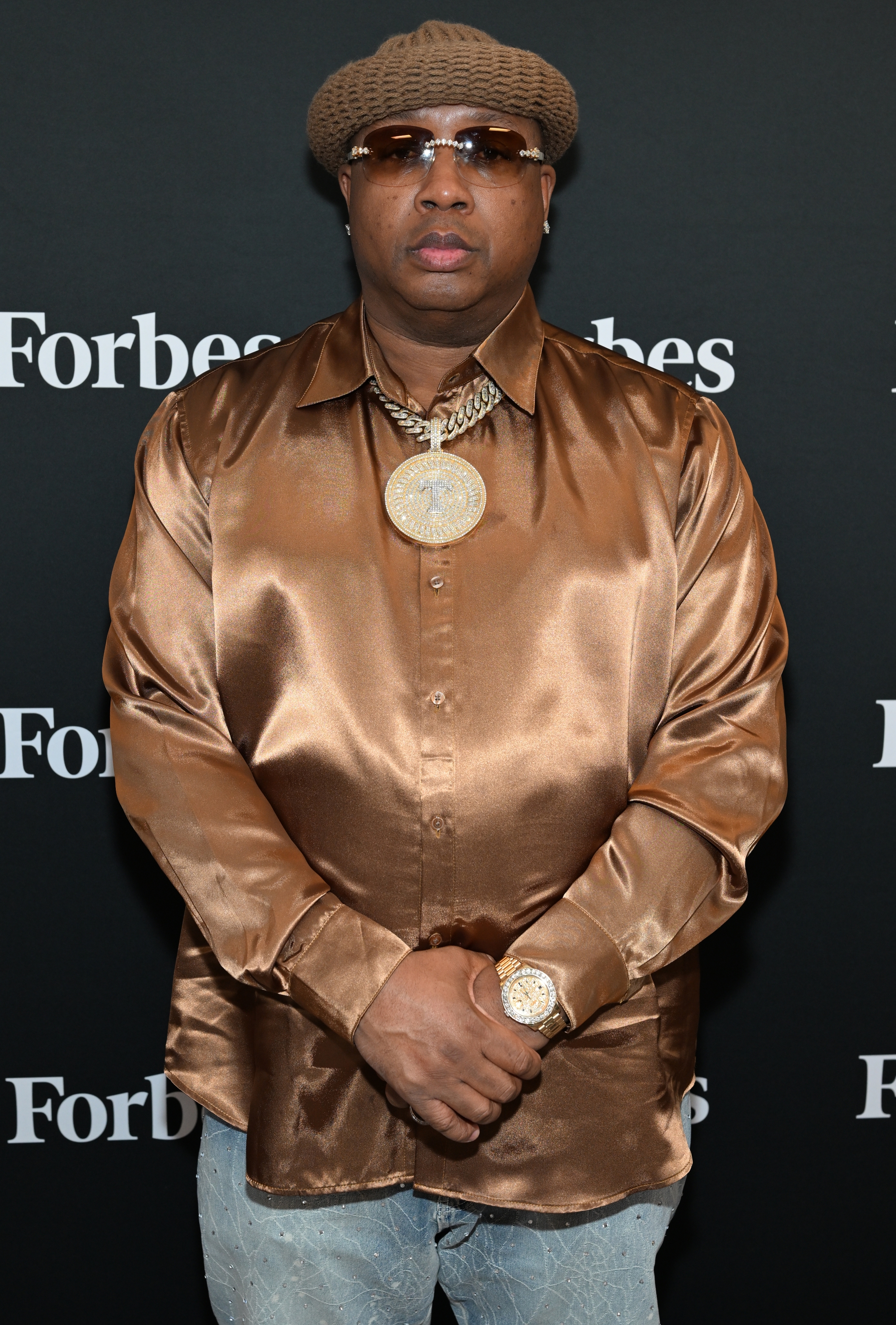Man wearing a shiny button-up shirt, ample  pendant necklace, and knit beret attends a Forbes event