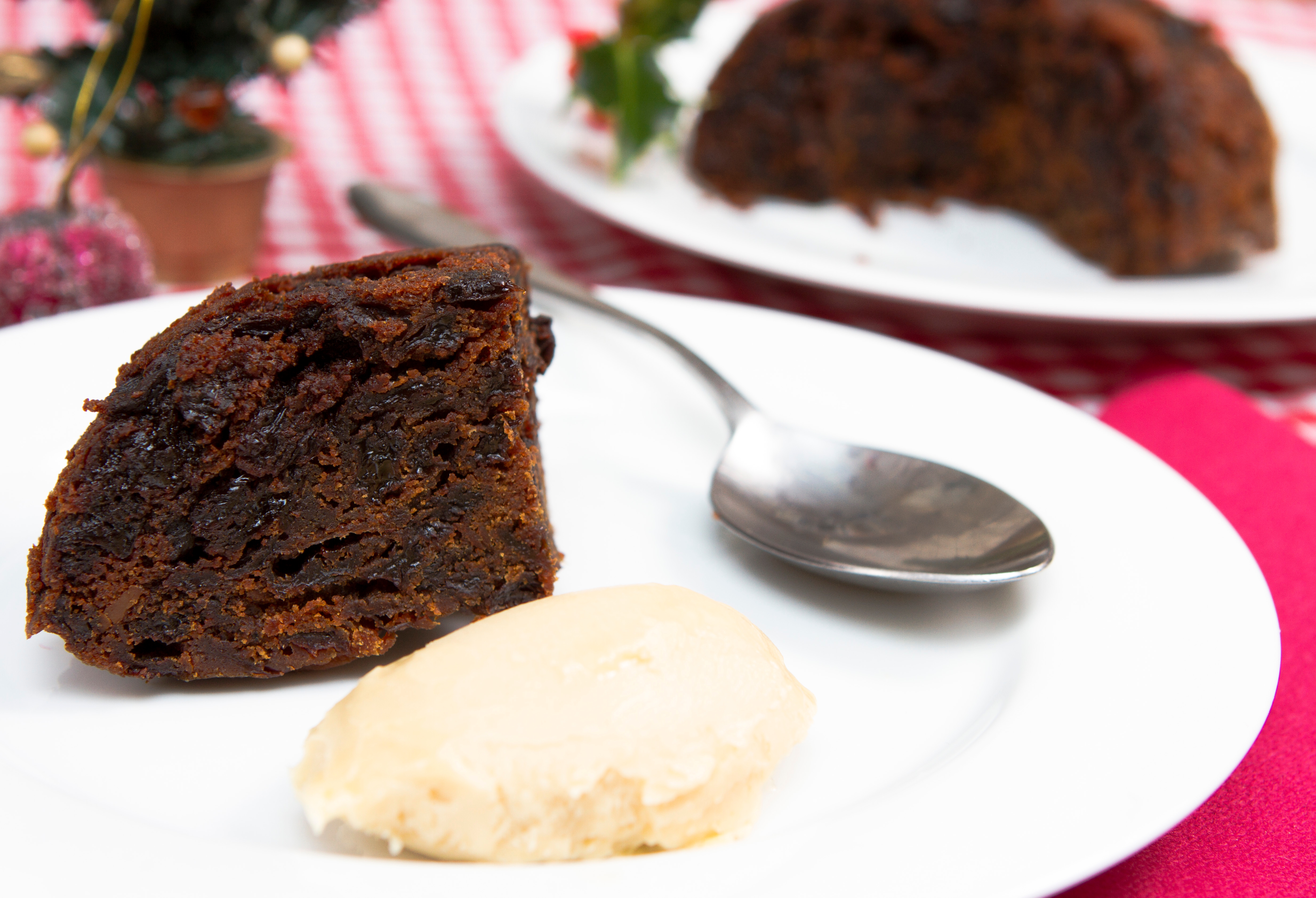 Slice of rich fruitcake on a plate with a dollop of cream, festive decor blurred in the background