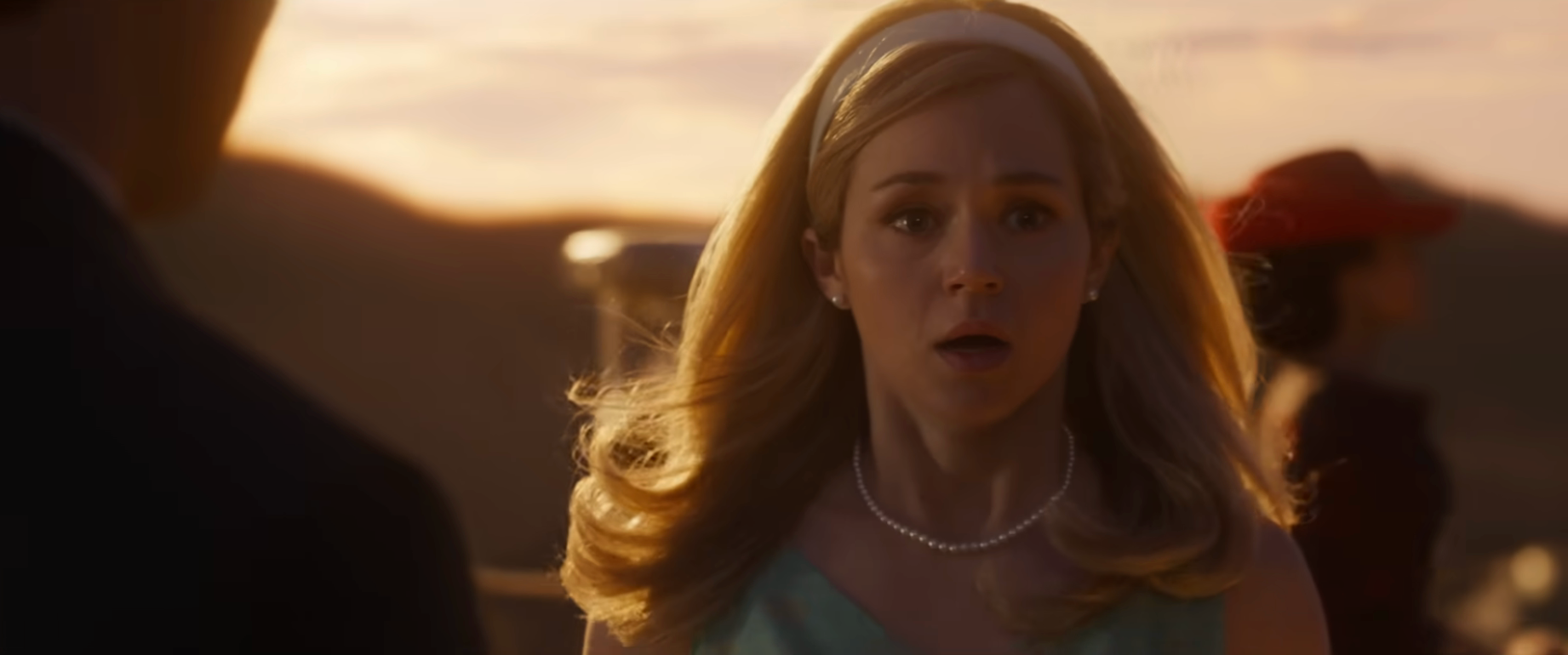 A pistillate   with agelong  hairsbreadth  looks surprised, wearing a pearl necklace and sleeveless dress, against a sunset backdrop with blurred figures