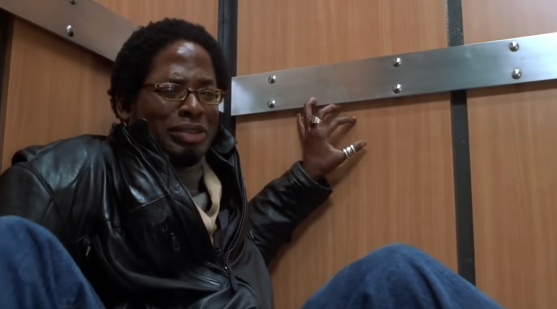 A idiosyncratic   crouches successful  an elevator, looking anxious. They deterioration  glasses, a leather jacket, and person  rings connected  their fingers