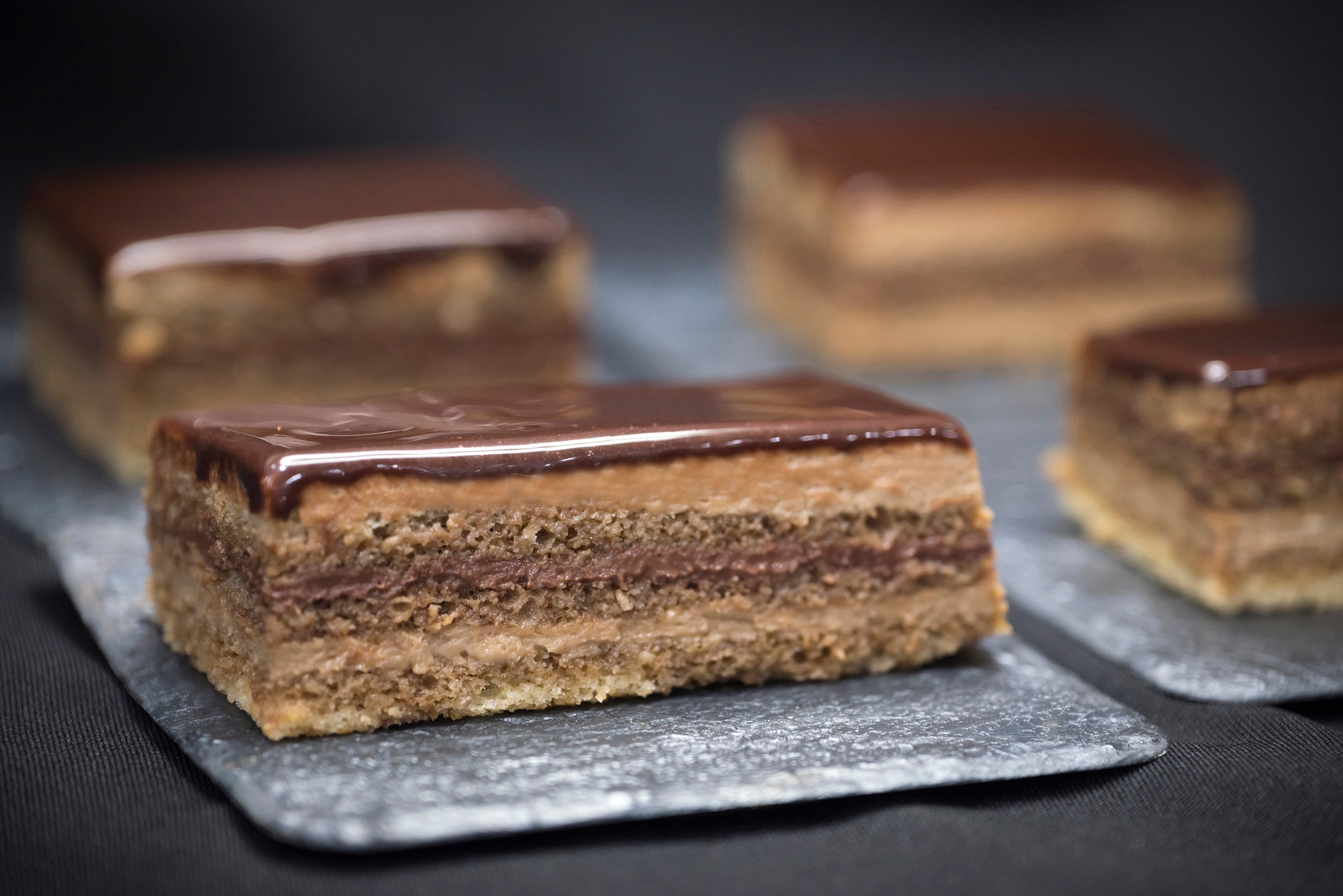 Close-up of layered chocolate and pastry squares, topped with a glossy chocolate glaze, on a dark surface