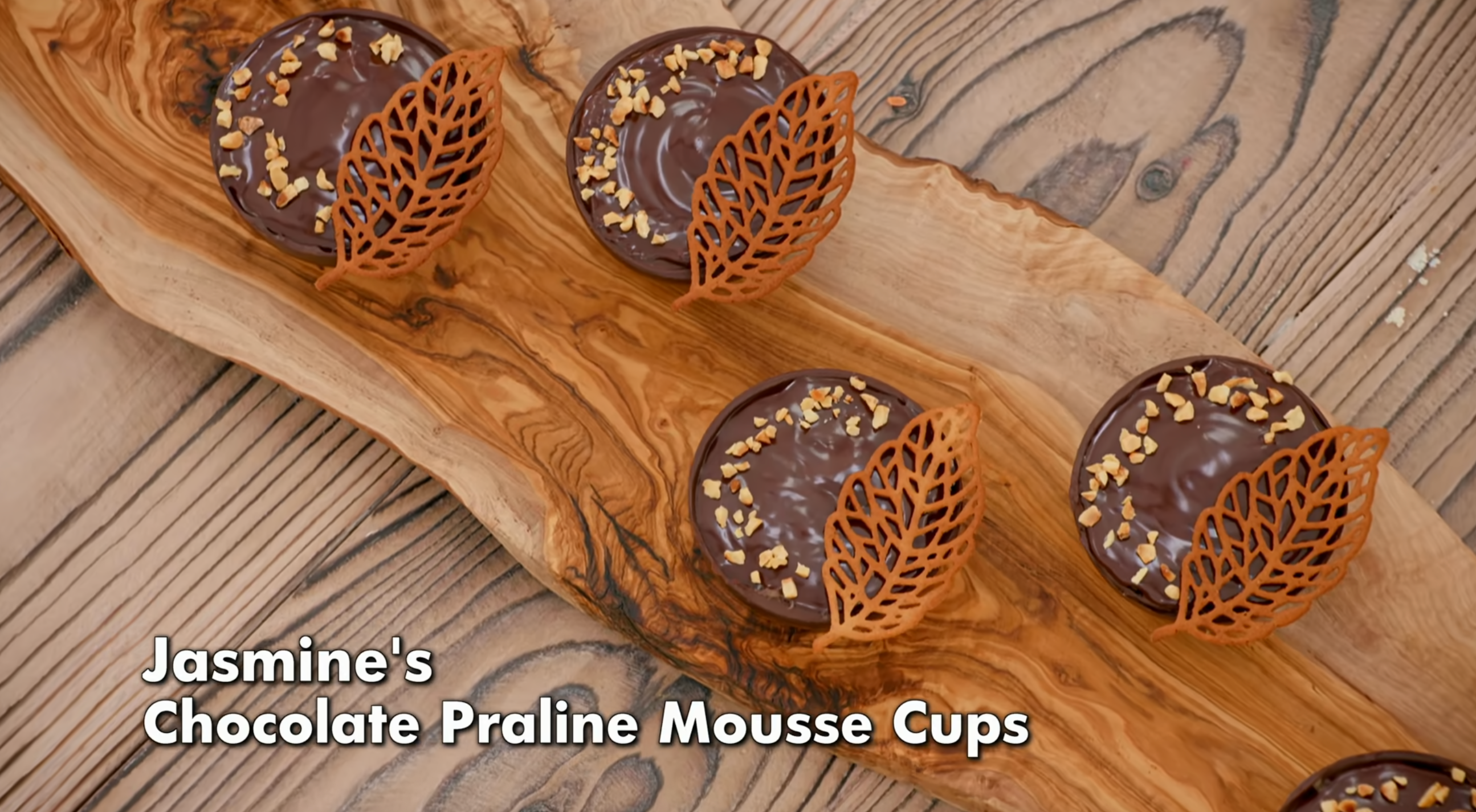 Chocolate praline mousse cups topped with caramelized nuts and decorative chocolate leaves, arranged on a wooden board. Text: &quot;Jasmine&#x27;s Chocolate Praline Mousse Cups.&quot;