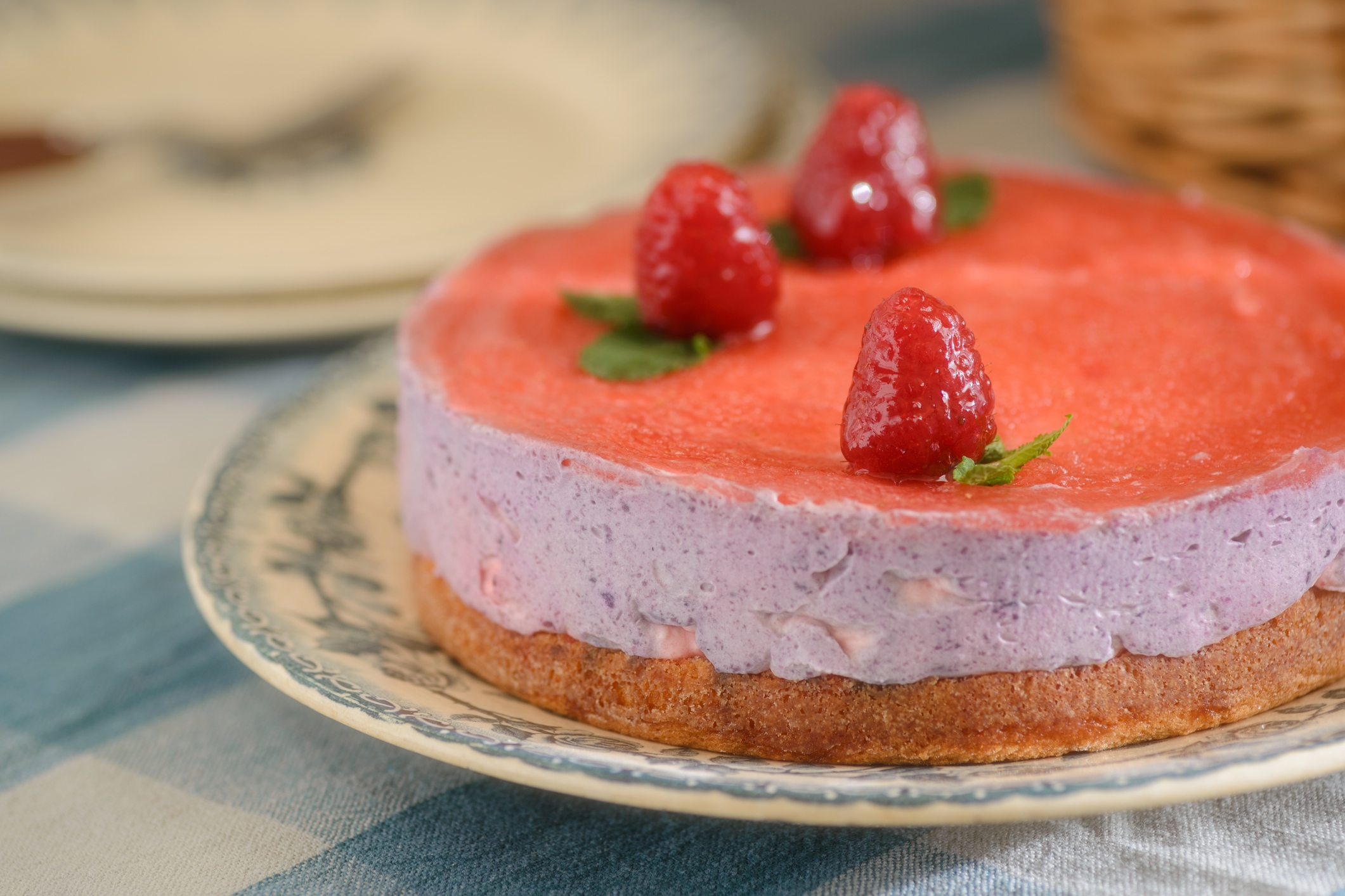 A cheesecake topped with a layer of strawberry glaze, fresh raspberries, and mint leaves on a decorative plate