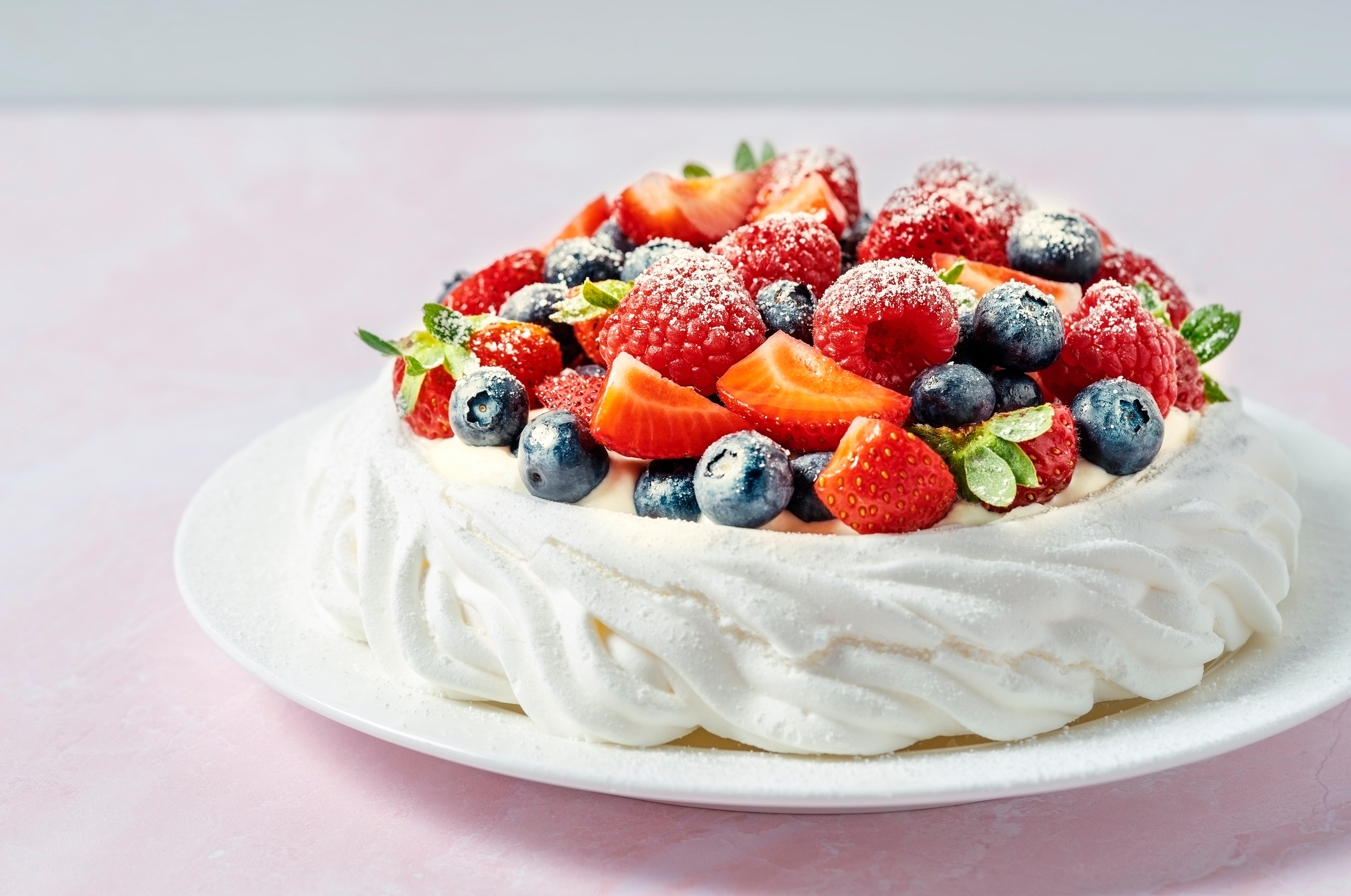 A pavlova topped with fresh strawberries, blueberries, and raspberries, dusted with powdered sugar, on a white plate