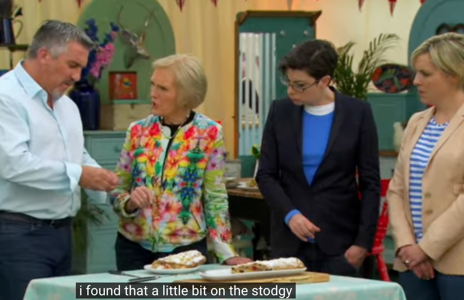 Four people discussing baked goods at a table on a cooking show set, with one person pointing at the pastries