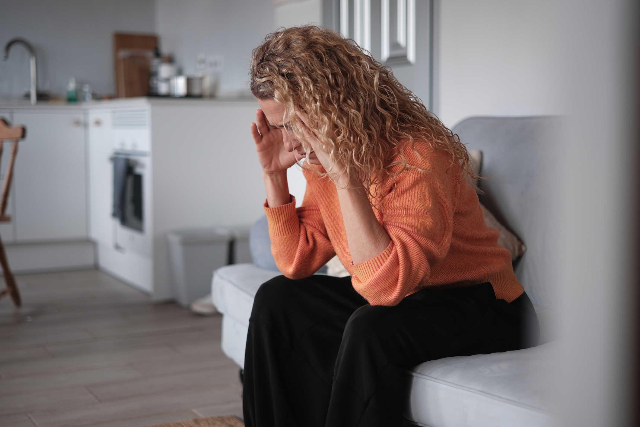 Person med krøllete hår som sitter i en sofa, holder hodet i hendene, virker stresset i hjemmemiljø