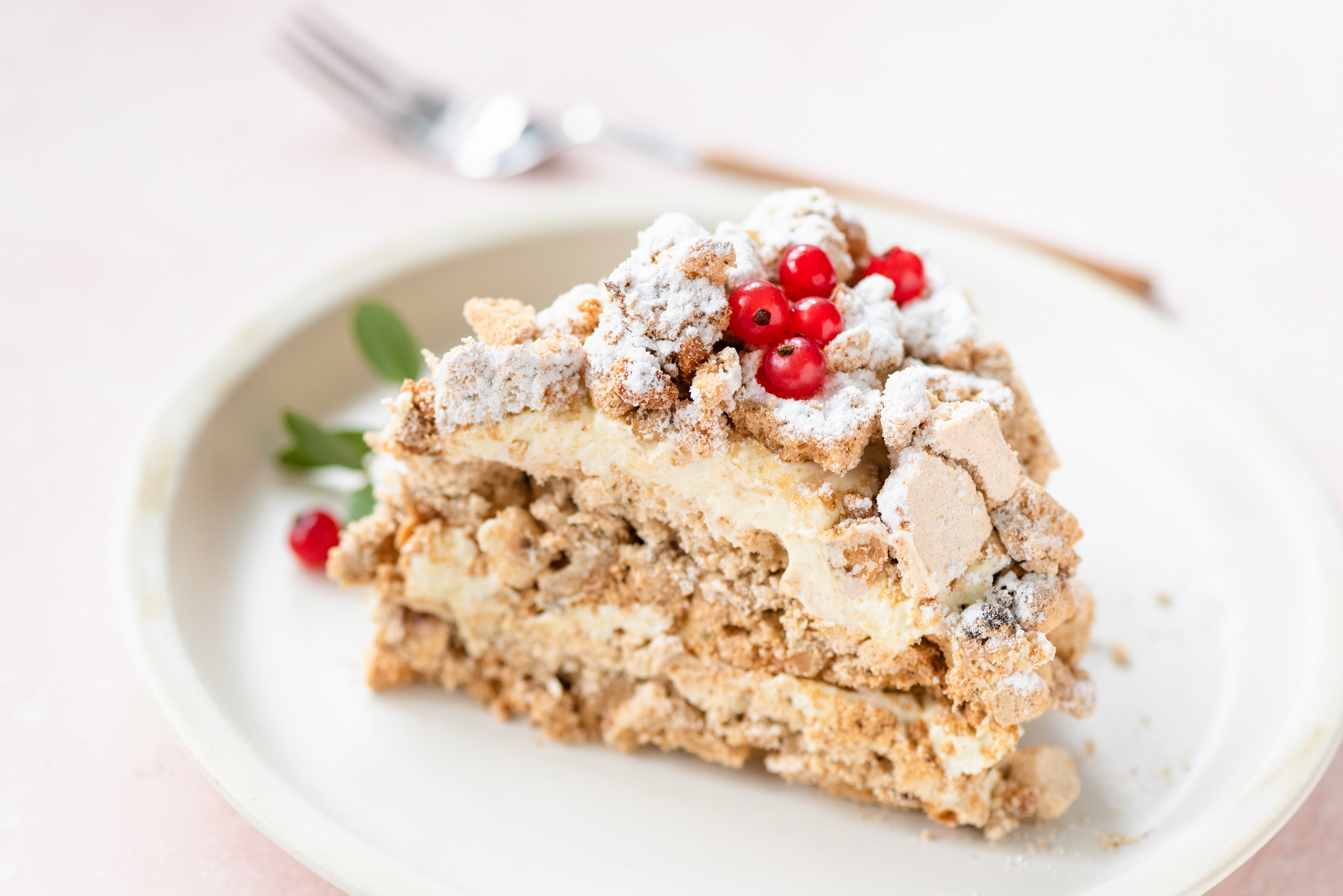 Slice of layered cake with cream filling, topped with crumbly pieces and red berries, on a white plate. Fork in the background