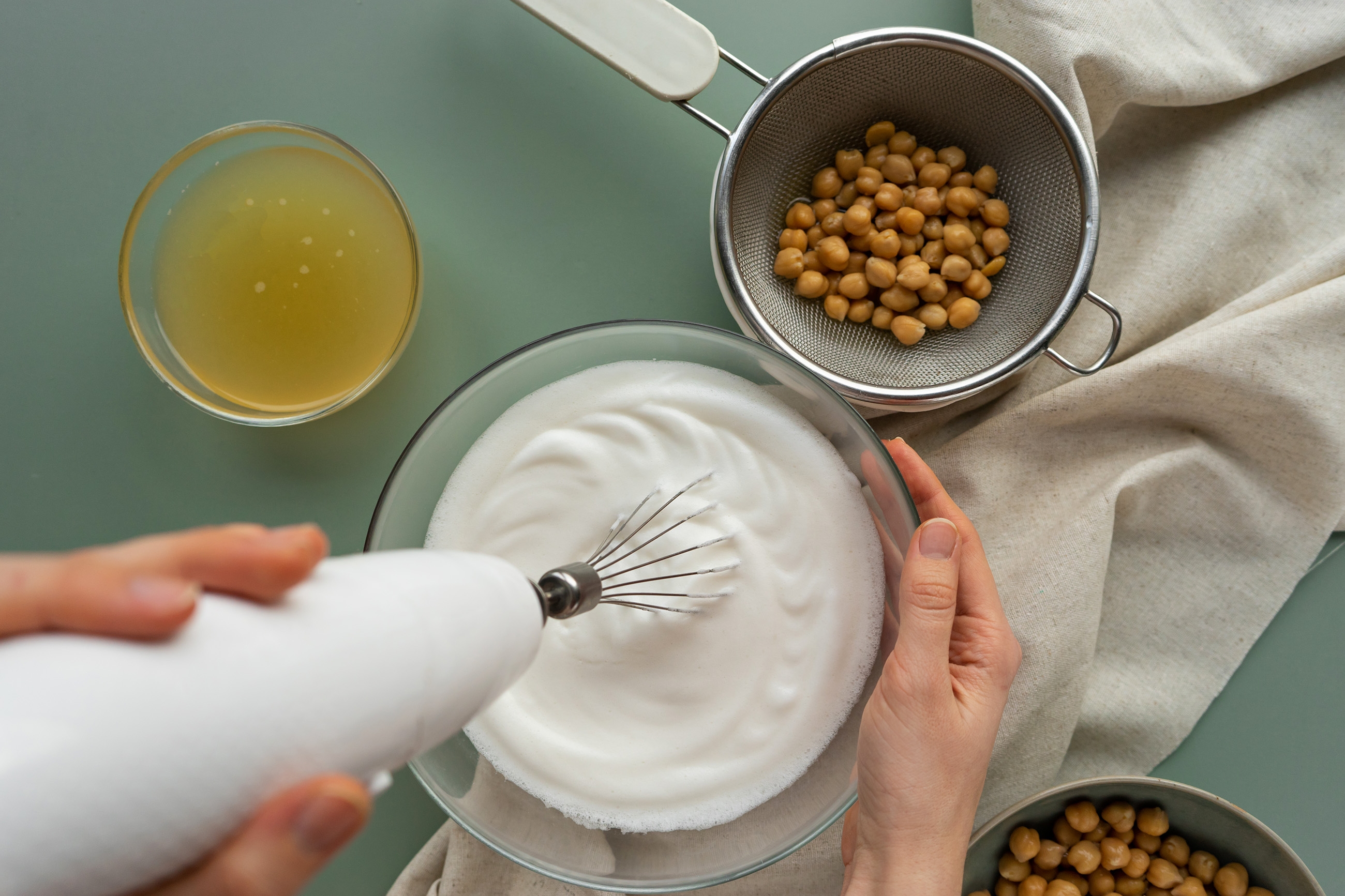 A person uses a hand mixer to whip chickpea liquid in a bowl, with chickpeas in a strainer and aquafaba in a glass nearby