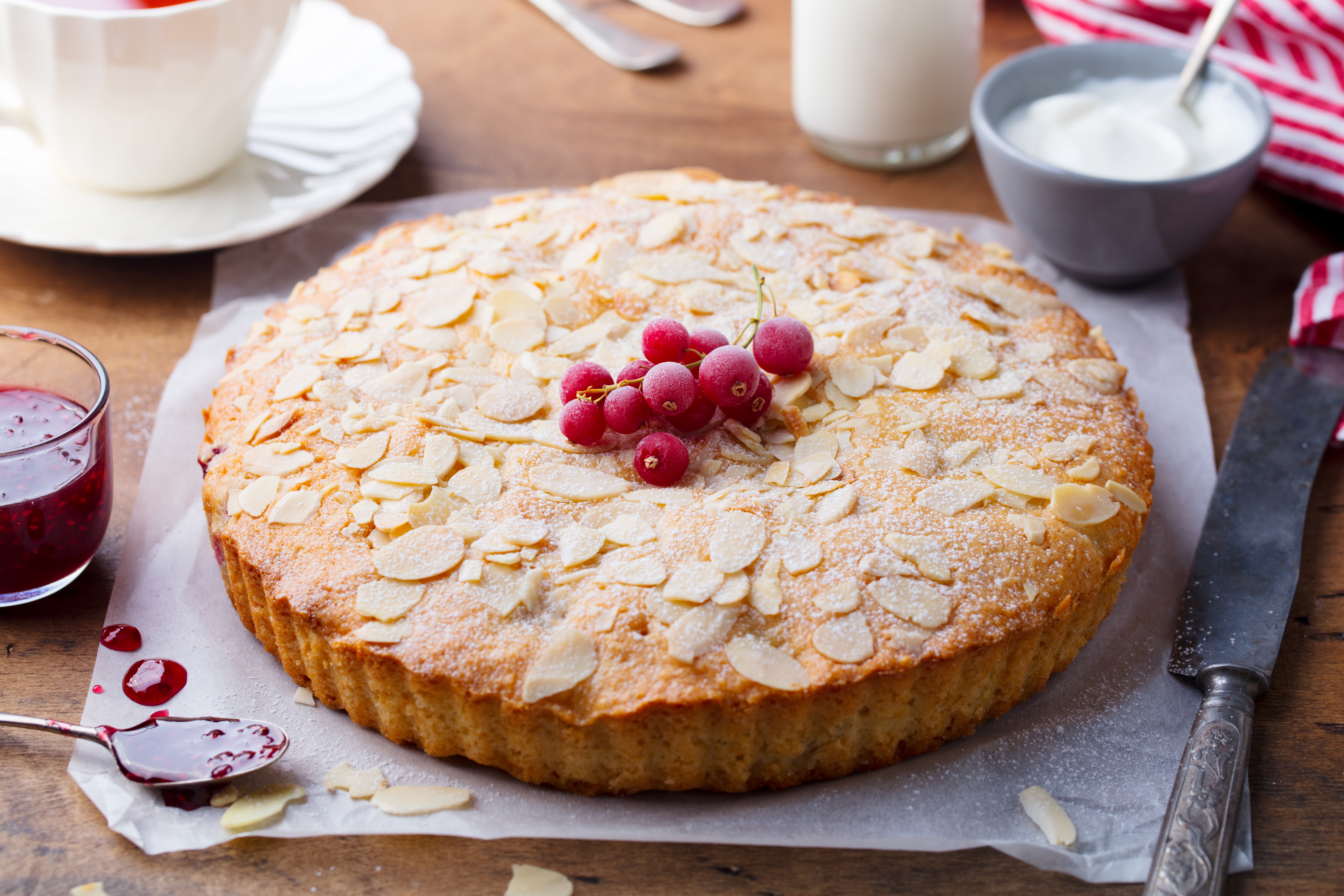 Almond cake topped with red currants on parchment paper, surrounded by a knife, berry sauce, and a dish of cream on a wooden table