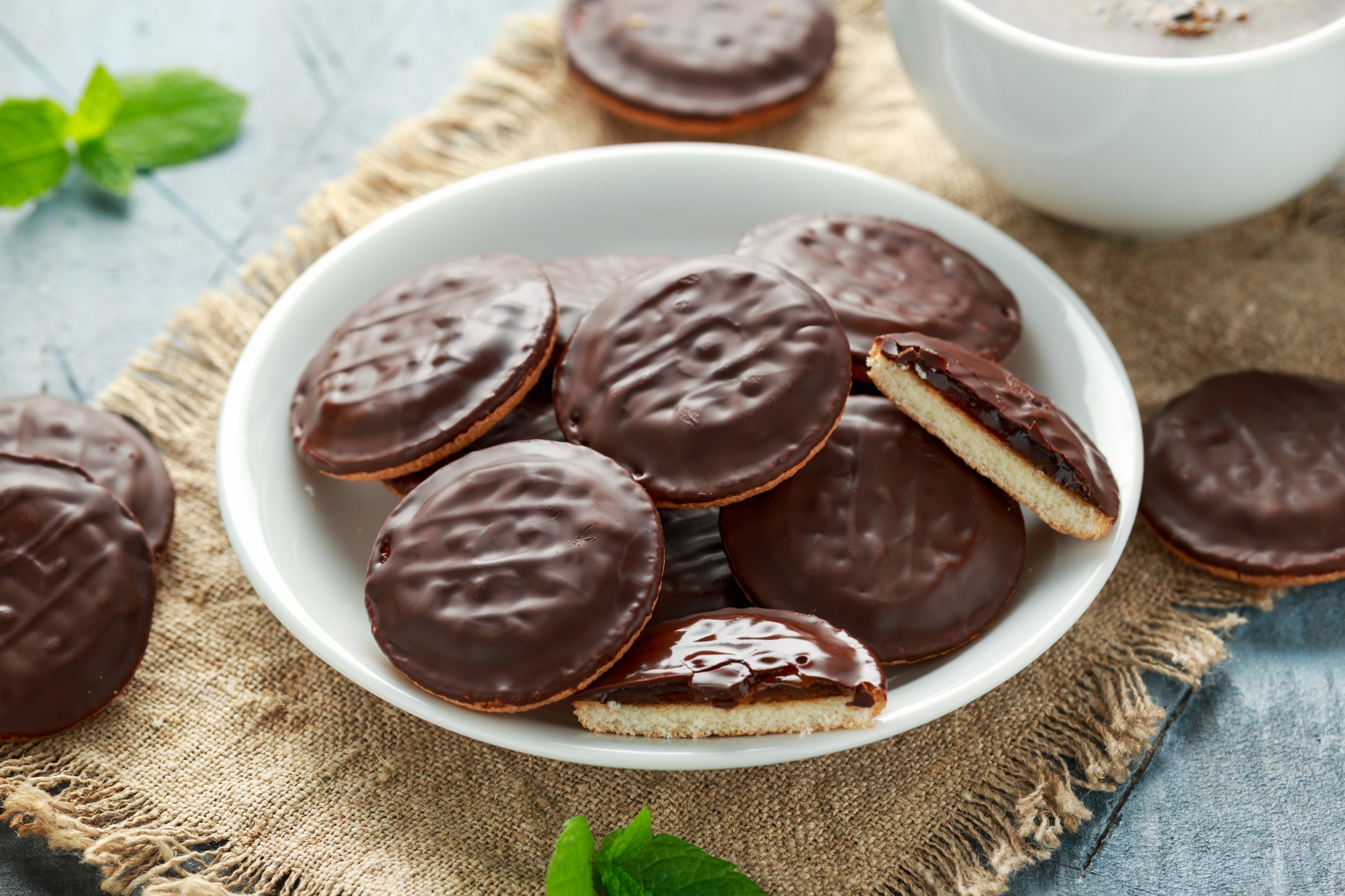 Plate of chocolate-covered biscuits on burlap placemat, with mint leaves scattered. A bowl of chocolate sauce is partially visible in the background