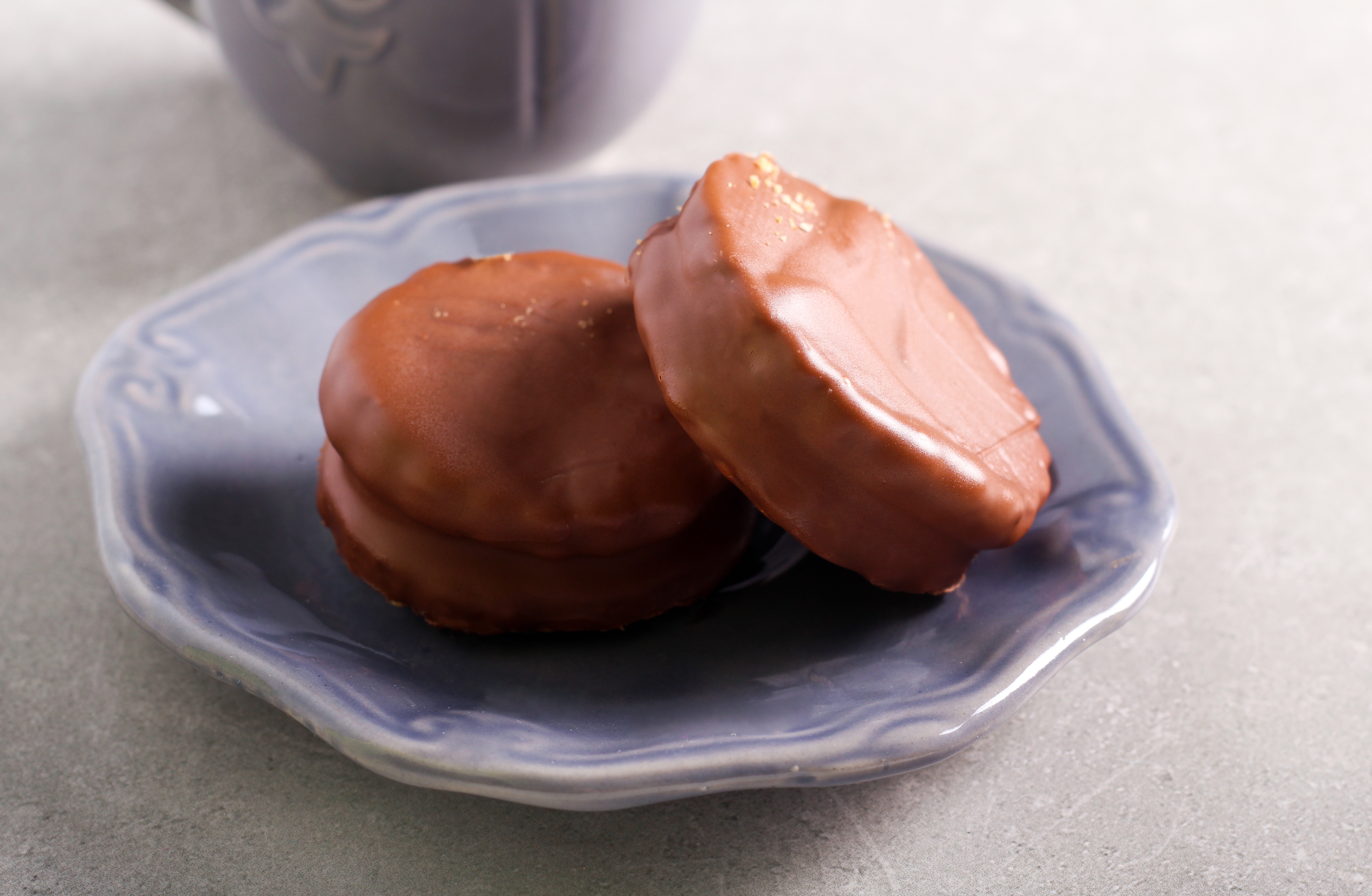 Two chocolate-covered cookies on a small plate with a mug partially visible in the background
