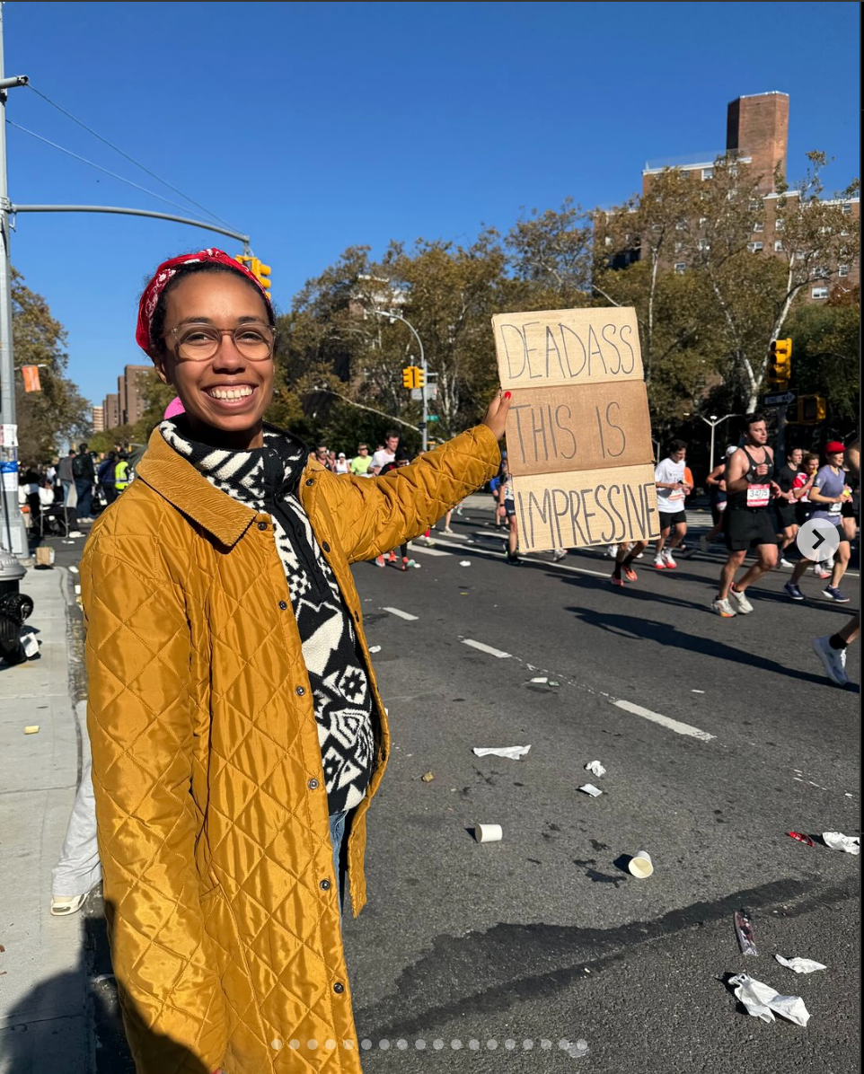 Person smiles and holds a sign reading "DEADASS THIS IS IMPRESSIVE" as runners pass by on a city street during a marathon