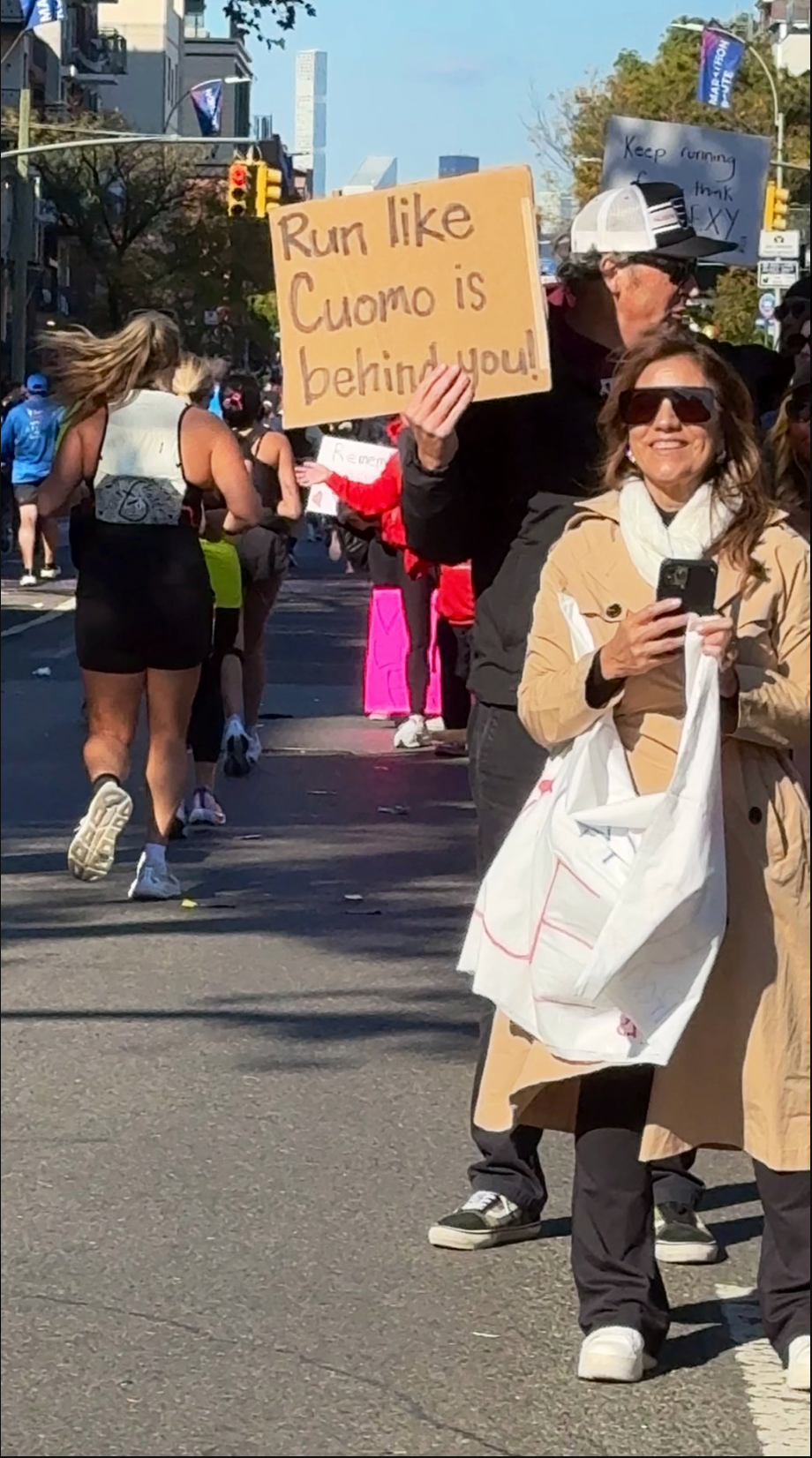 Two people watch a street run; one holds a sign that reads "Run like Cuomo is behind you!"