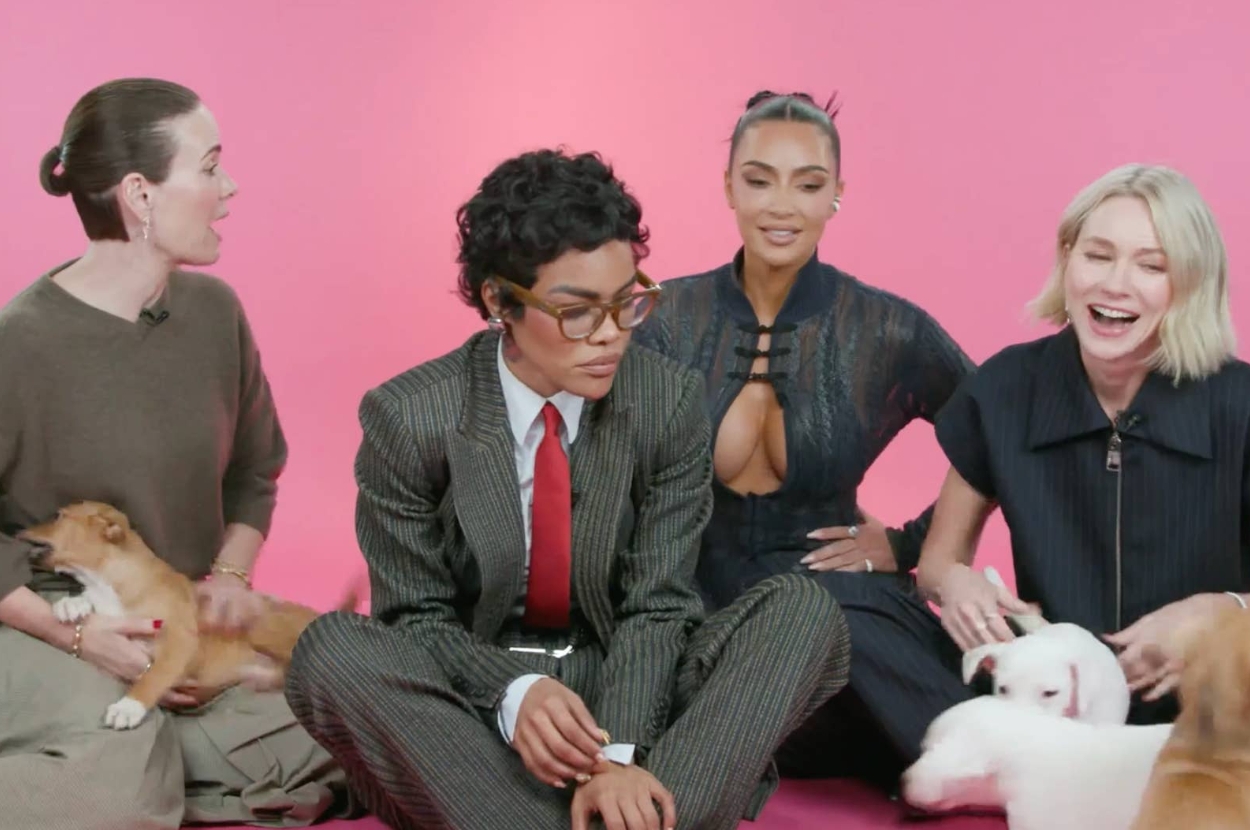 Group of four women, one in a pinstripe suit and tie, another in a cut-out lace outfit, with puppies, against a plain backdrop