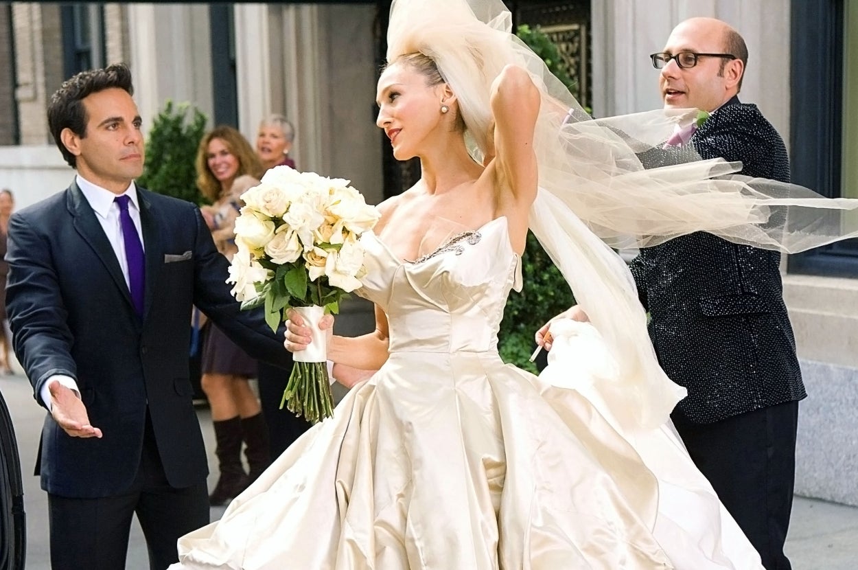 A person in a vintage-style wedding dress with a bouquet is assisted by two individuals on a city street, with her veil flowing behind her