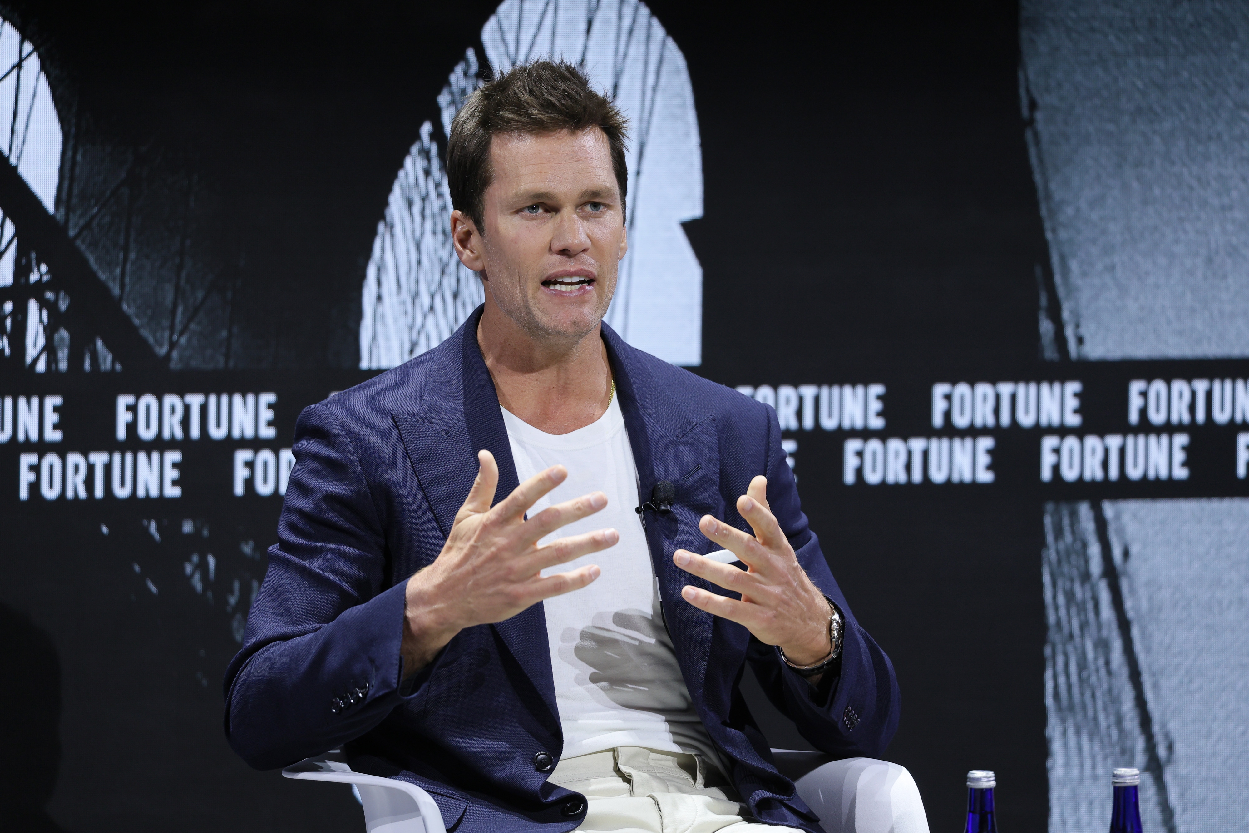 Tom Brady speaks energetically on stage at a Fortune event, wearing a casual blazer over a t-shirt while seated