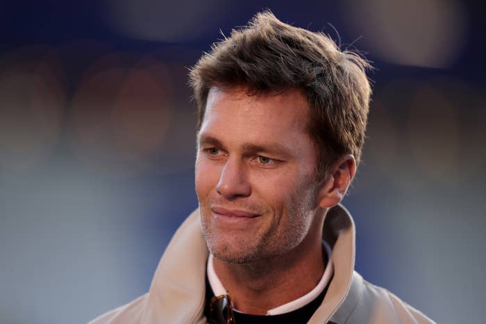Tom Brady in a jacket with short hair smiling slightly, outdoors in a blurred background