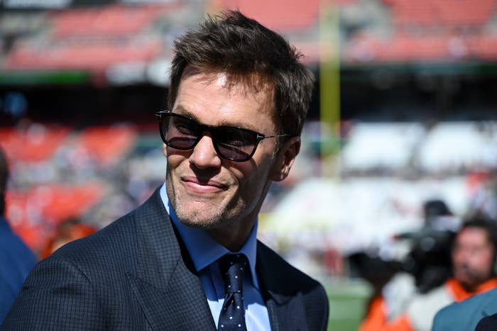 Tom Brady in a suit and tie, wearing sunglasses, smiles slightly while standing outdoors in a stadium setting
