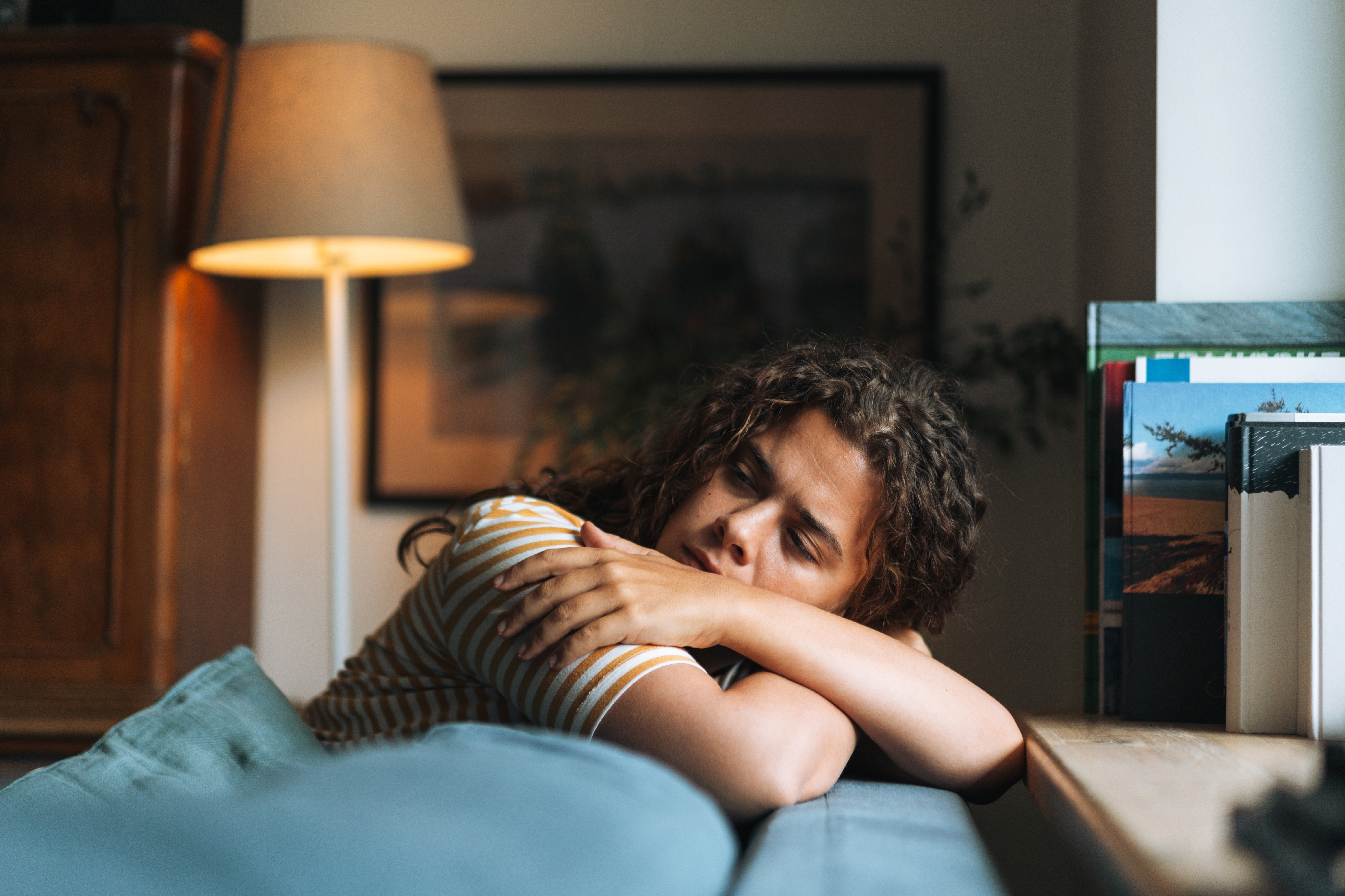 Person med krøllete hår som hviler hodet på foldede armer, ser fredelig og kontemplativ ut på en sofa i et koselig rom