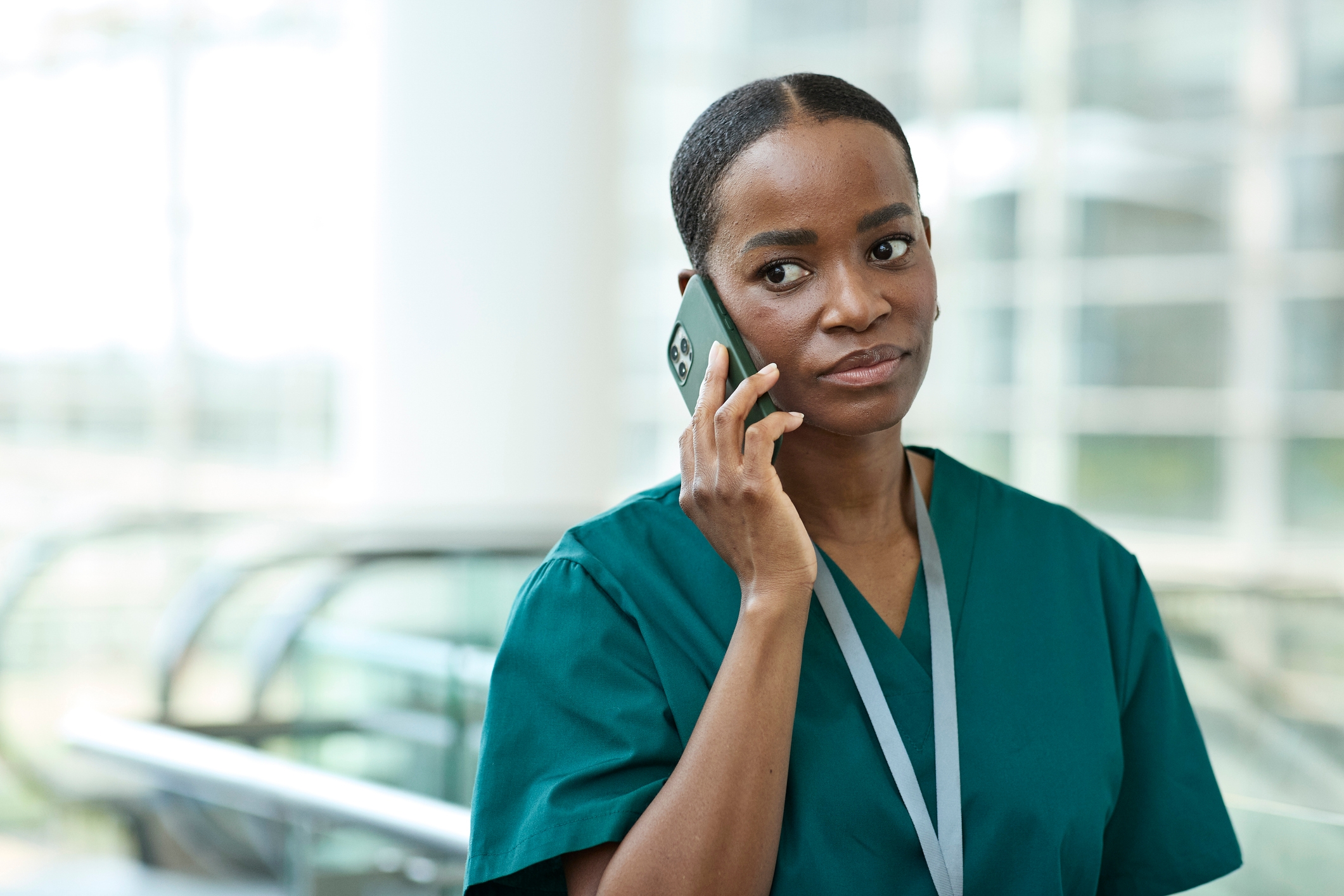 En person i grønn skrubb snakker i en mobiltelefon, står innendørs med en rulletrapp i bakgrunnen