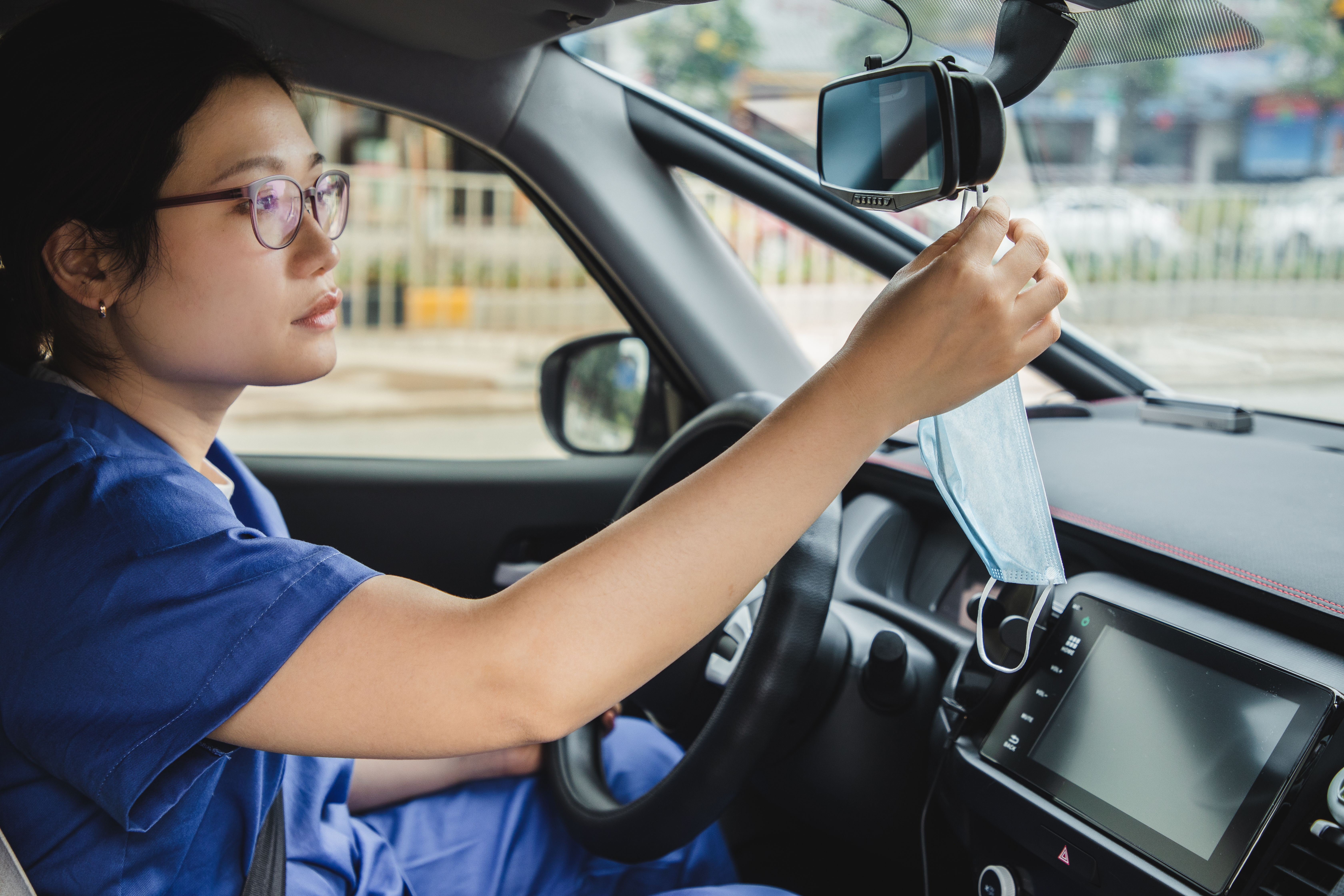 Person i scrubs henger en ansiktsmaske på en bils bakspeil, noe som antyder oppmerksomhet på helse og sikkerhet under pendling
