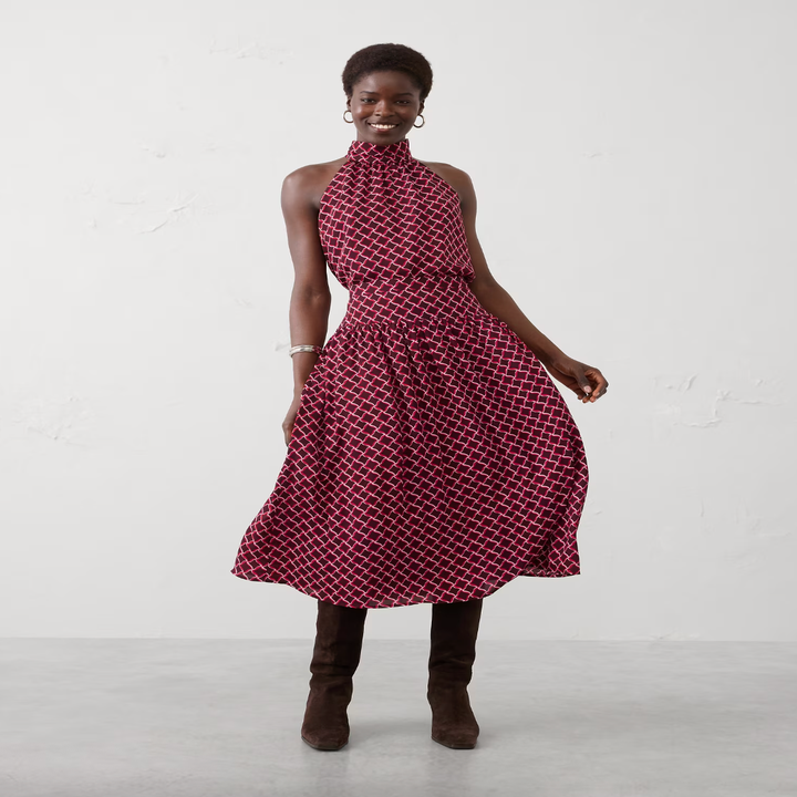 Person wearing a sleeveless, patterned dress with a high neck and skirt and tall boots, posing indoors