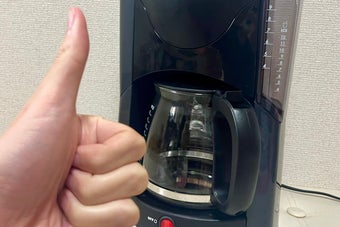 コーヒーメーカーの前で親指を立てる手。コーヒーマシンは電源が入っている状態。