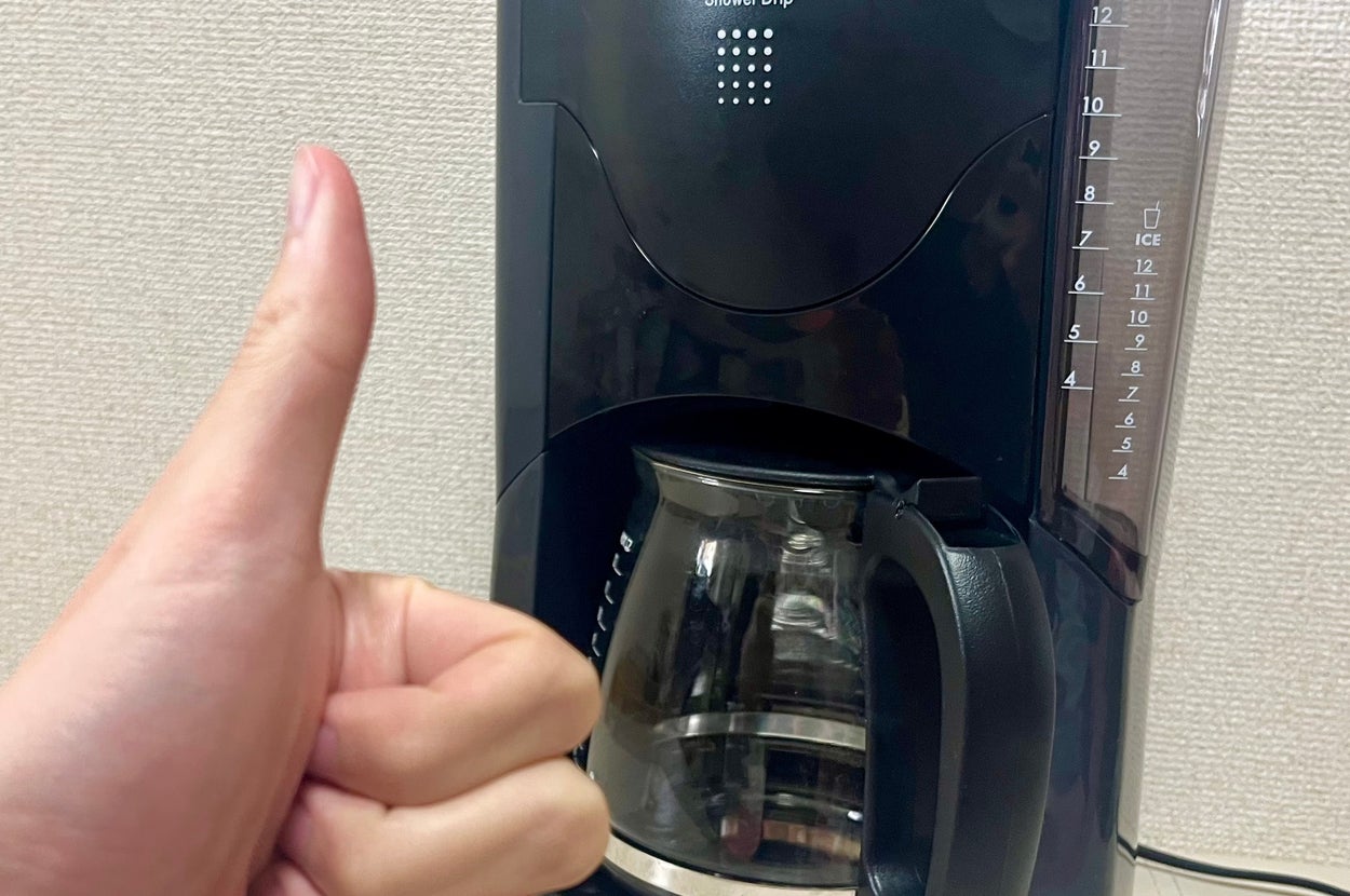 コーヒーメーカーの前で親指を立てる手。コーヒーマシンは電源が入っている状態。