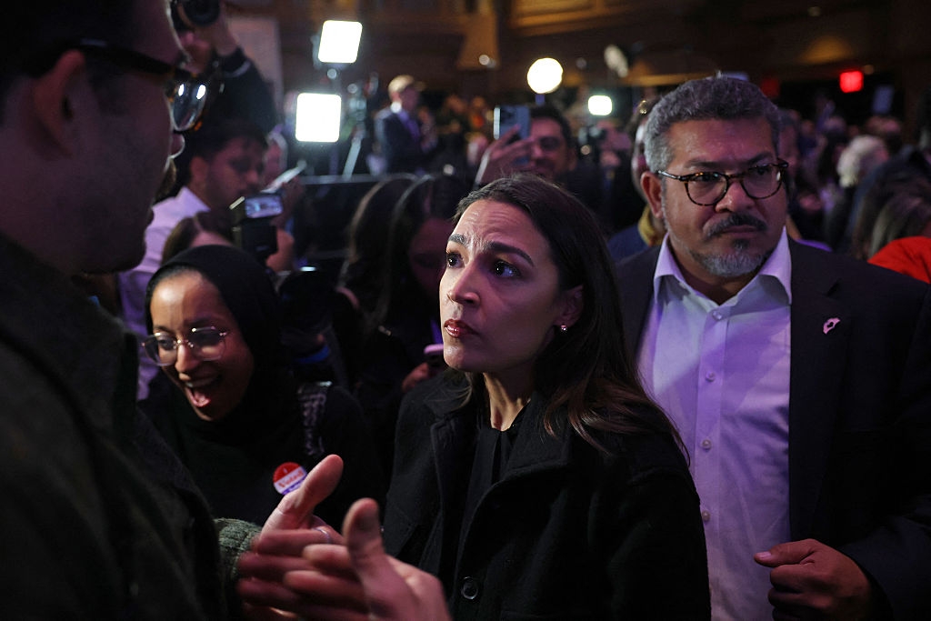 A woman in a black coat speaks to a man in a busy indoor setting, surrounded by people and photographers