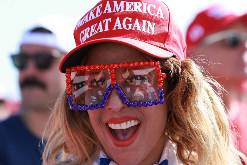 Person som har på seg en "Make America Great Again" hatt og store, utsmykkede solbriller, smiler bredt på et utendørsarrangement