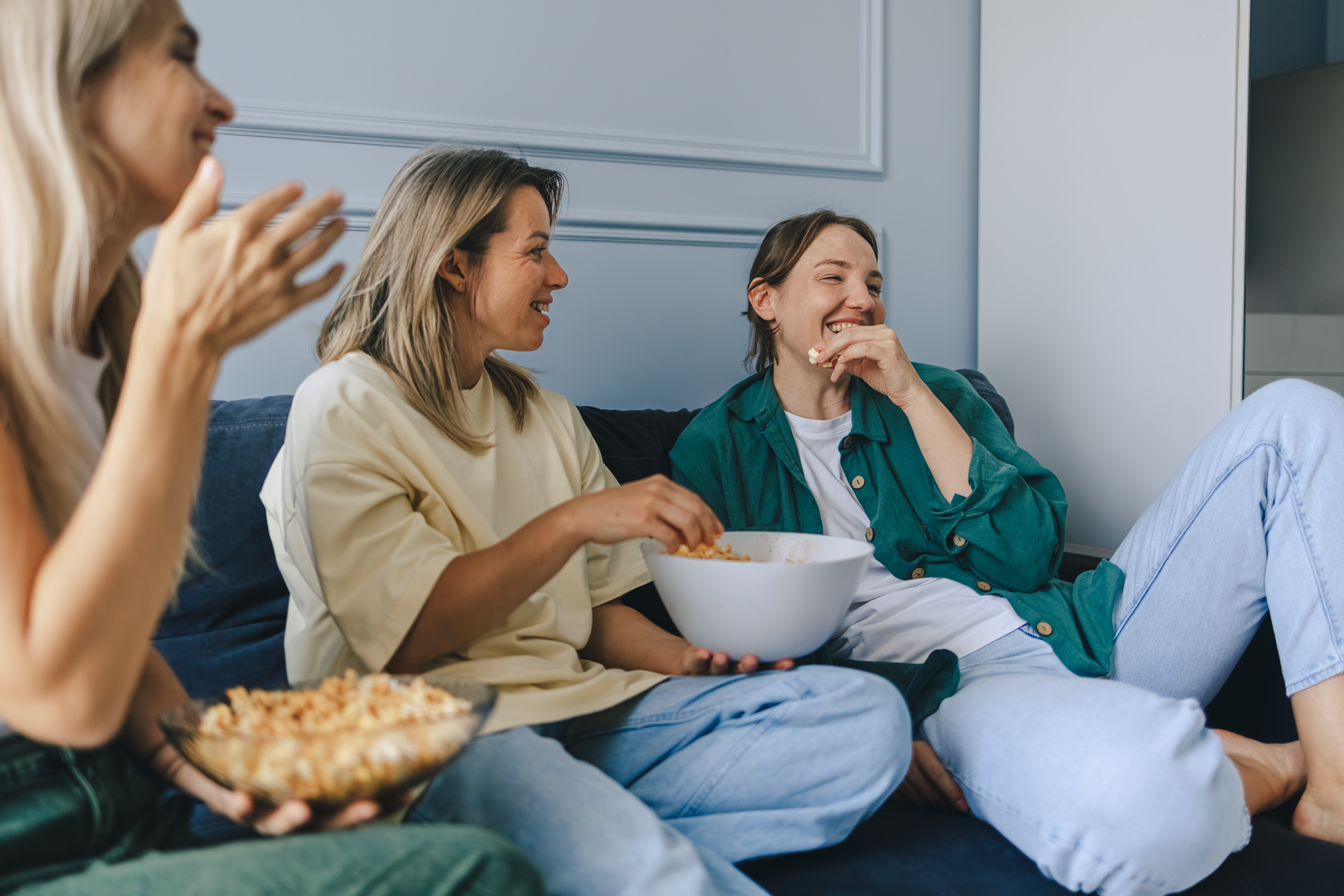 Tre personer sitter på en sofa, smiler og spiser snacks fra boller, og deler et uformelt, gledelig øyeblikk sammen