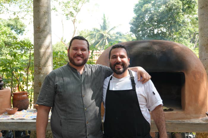 Two people smiling in front of an outdoor brick oven. Both appear relaxed and happy, wearing casual and chef attire