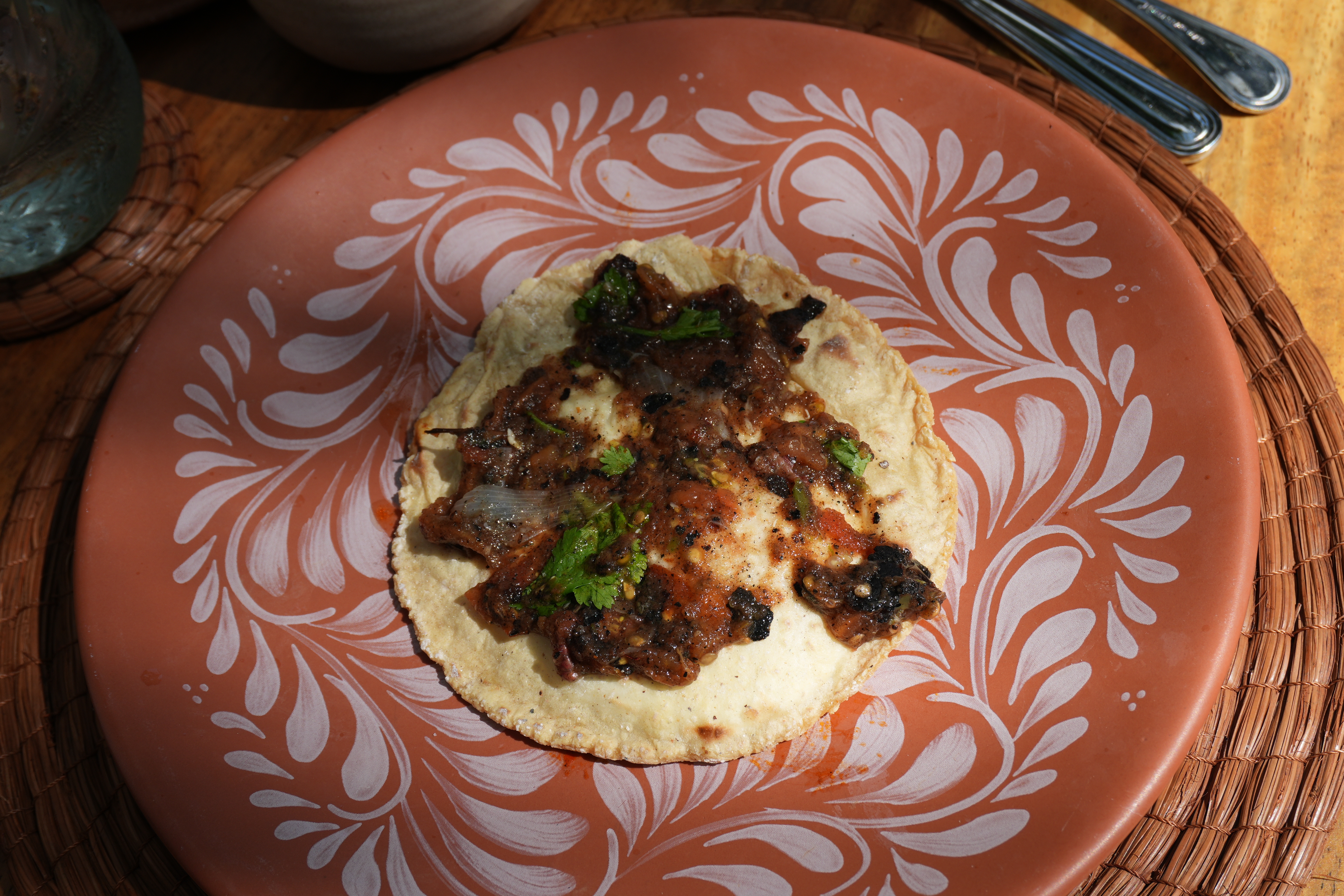 Taco with charred meat and herbs on a decorative plate, placed on a woven mat beside cutlery and a bowl