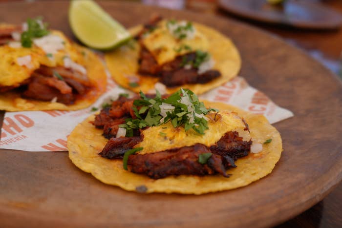 Three tacos with grilled meat, onions, and cilantro on a wooden plate, served with a lime wedge