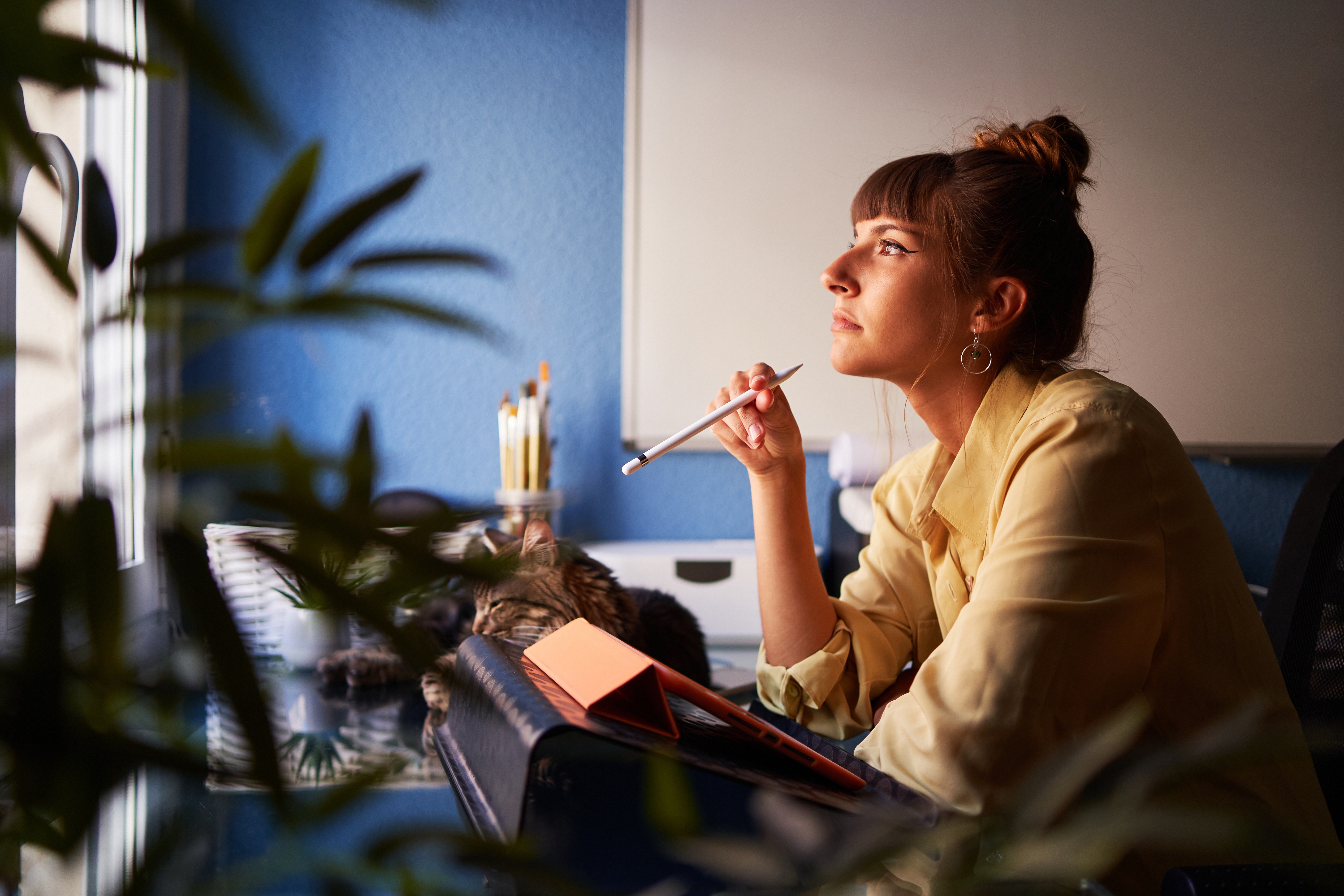 Kvinne i en gul bluse sitter ved et skrivebord, holder en penn mot haken og ser ettertenksomt ut av vinduet