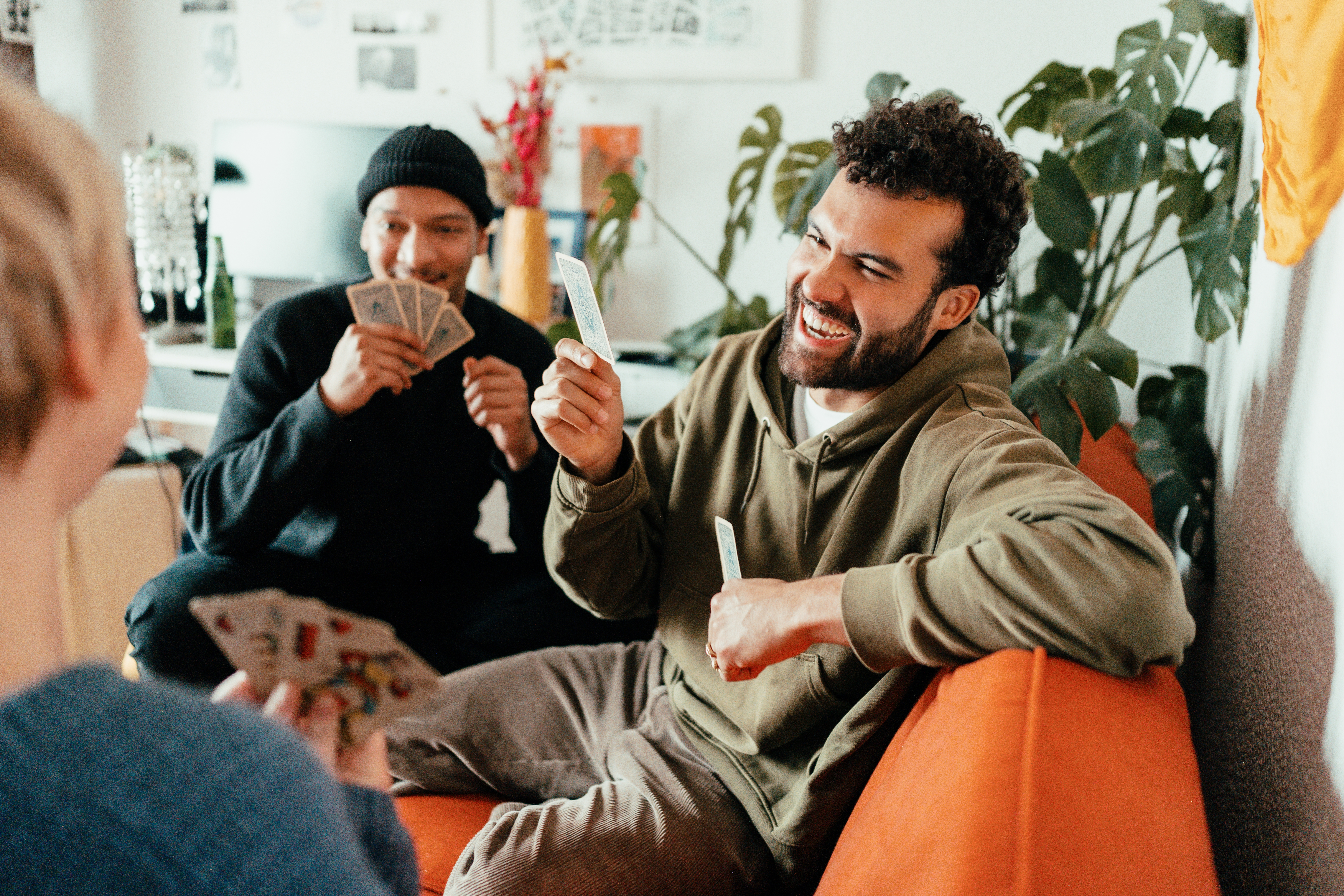 Tre personer sitter i en sofa og spiller kort og ler i en koselig stue