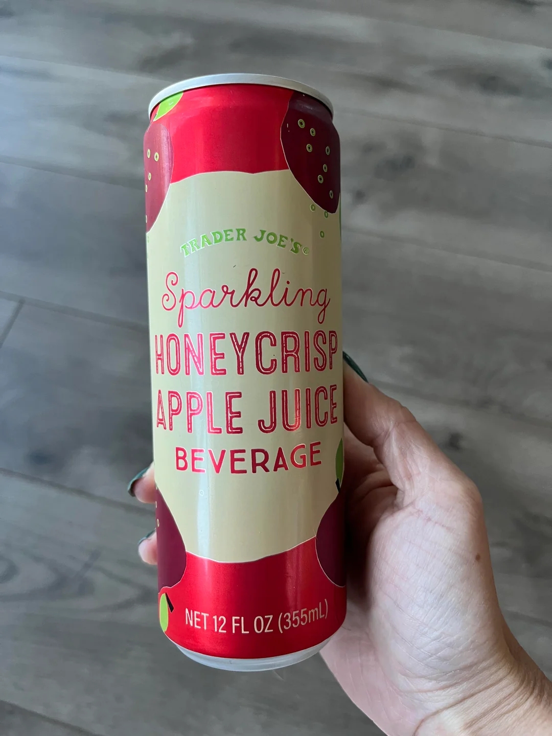 A hand holds a can of Trader Joe&#x27;s Sparkling Honeycrisp Apple Juice Beverage on a wooden floor background