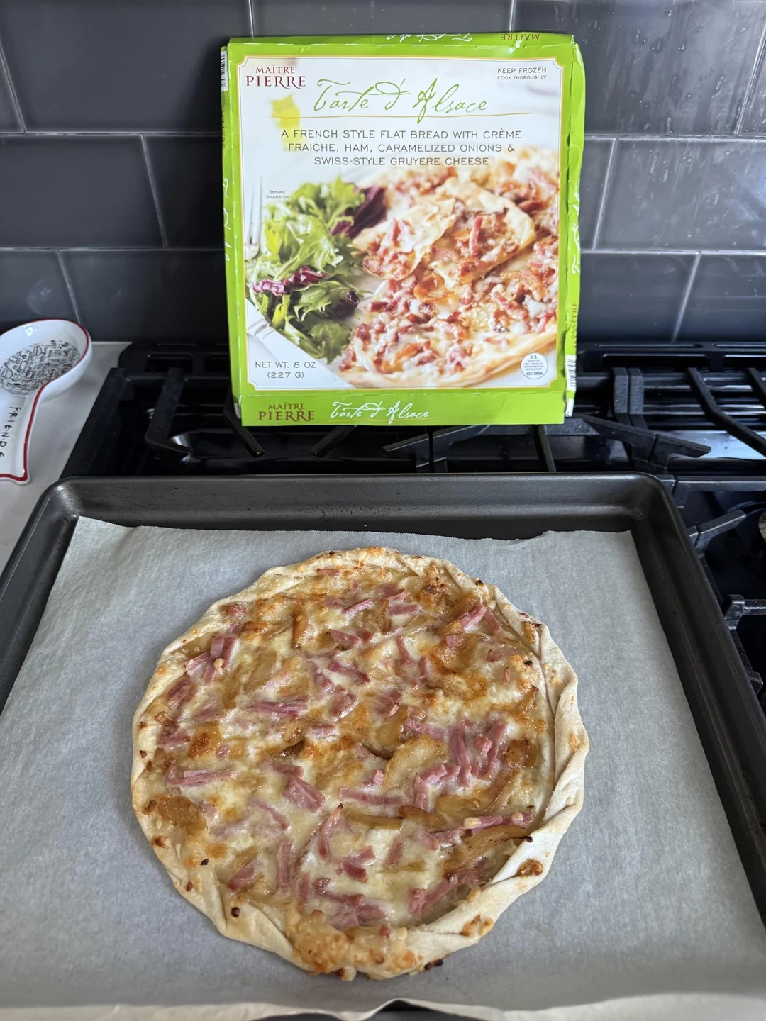 Frozen French-style flatbread with ham, caramelized onions, and cheese on a baking tray. A package with similar design is visible in the background