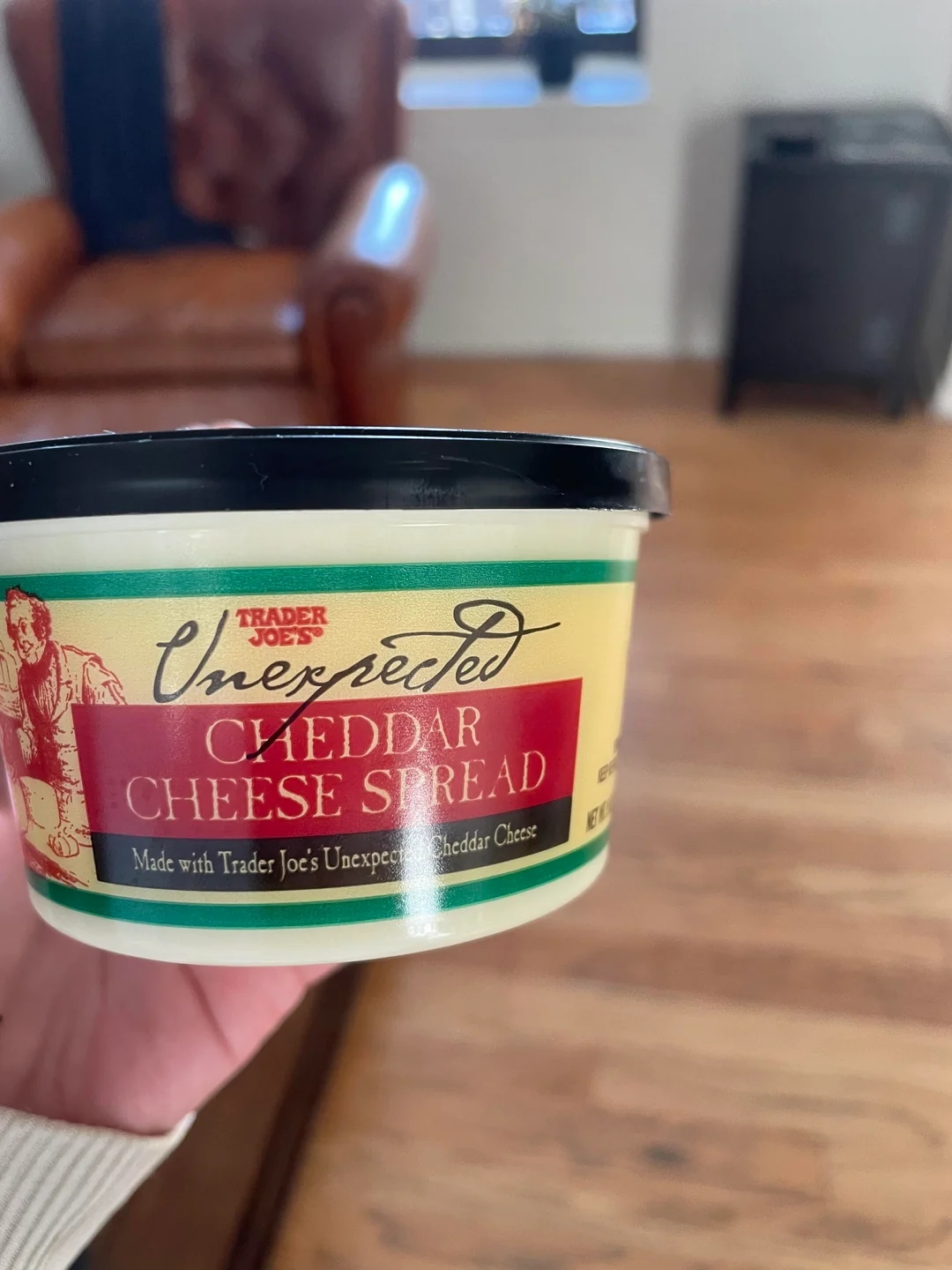 Hand holding a container of Trader Joe&#x27;s Unexpected Cheddar Cheese Spread, with a red armchair in the background