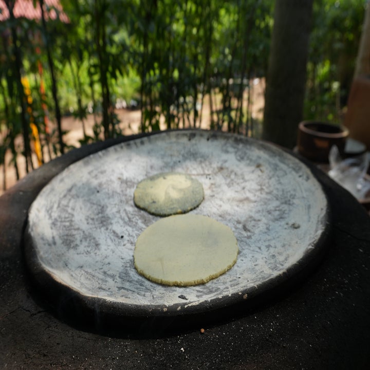 Two tortillas cooking on a large traditional stone griddle outdoors, surrounded by a lush garden setting