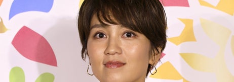 ショートカットの女性が白いシャツと赤いエプロンを着て、背景に抽象的な花柄がある。