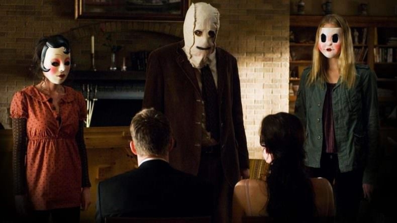 Three people in eerie masks stand menacingly before two seated individuals in a dimly lit room, creating a tense atmosphere