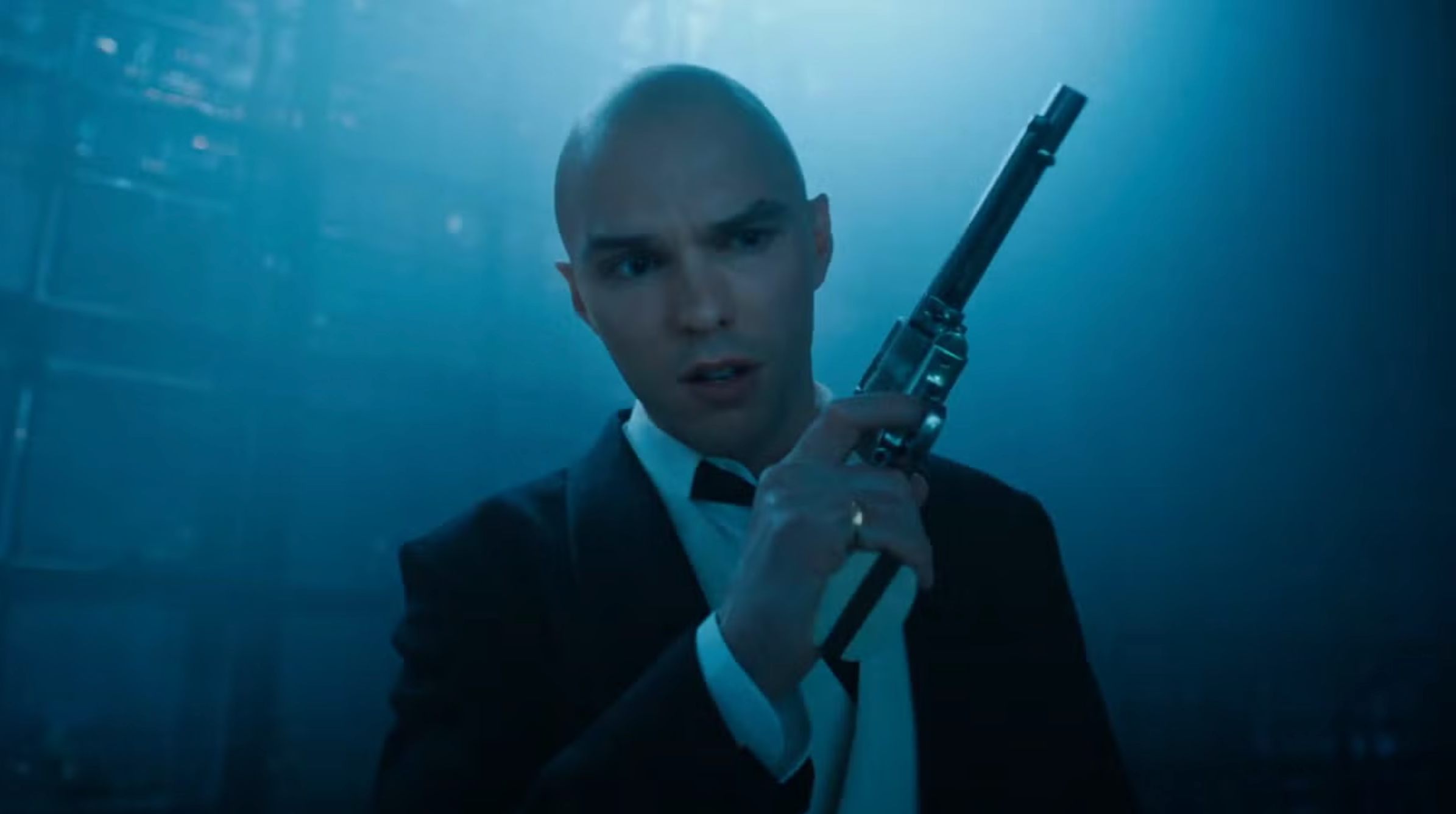 Person in a suit holds a gun, looking serious in a dimly lit setting