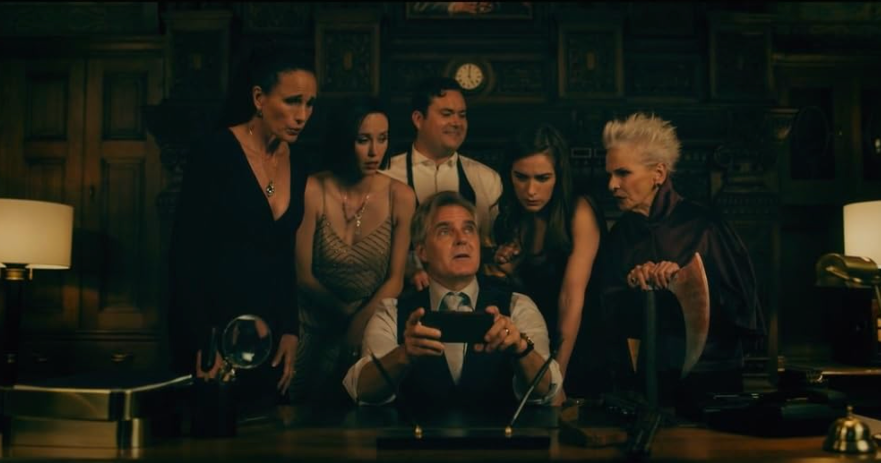 Group of six people in stylish, dramatic attire gather intently around a man sitting at a desk with a focused expression, holding a small object