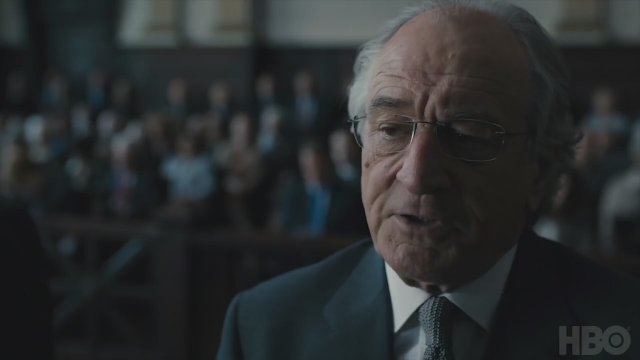 An older man in a suit stands in a courtroom setting, speaking, with people seated in the background