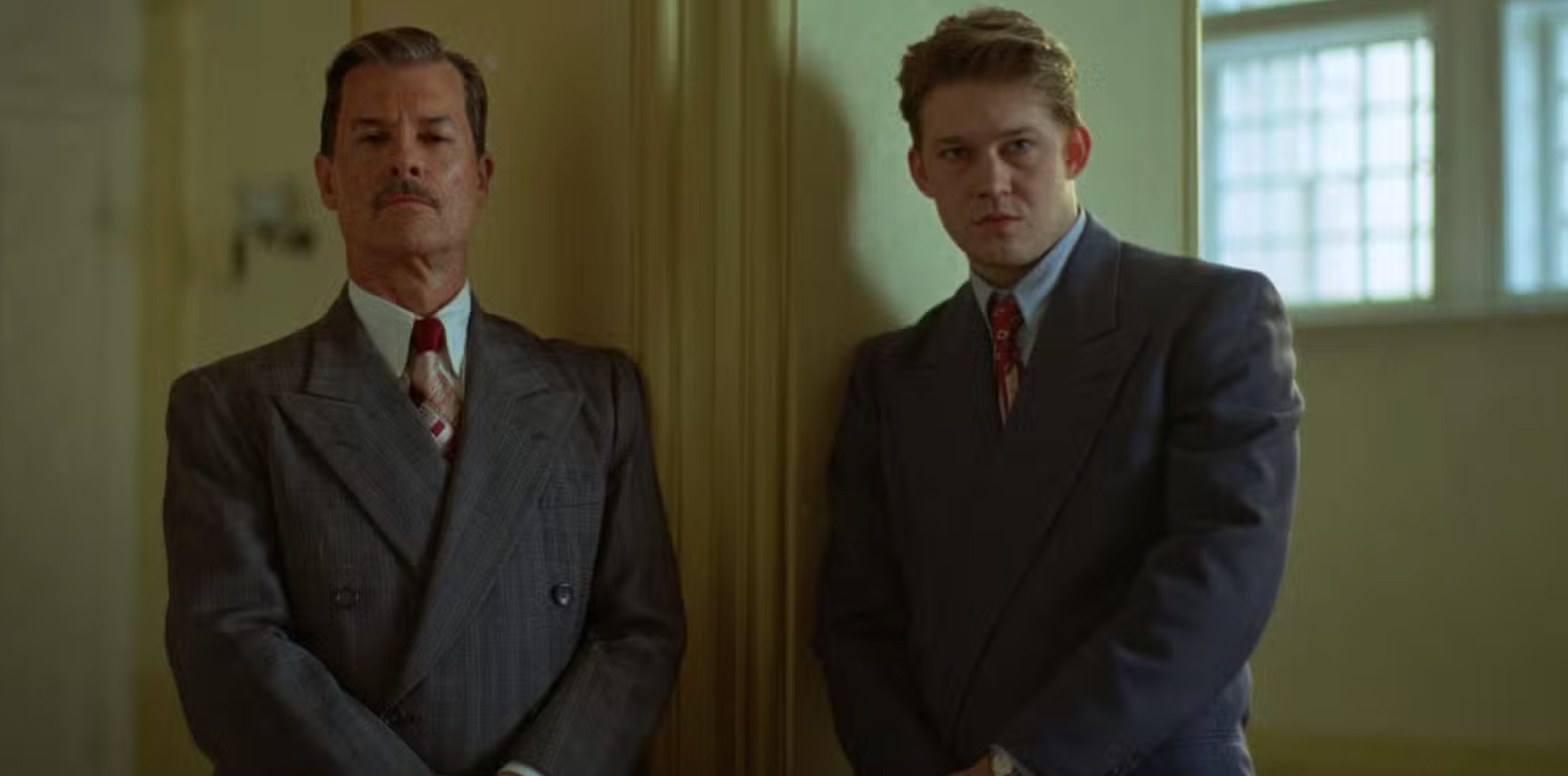 Two people in vintage-style suits stand against a wall, looking serious. The setting appears formal or official