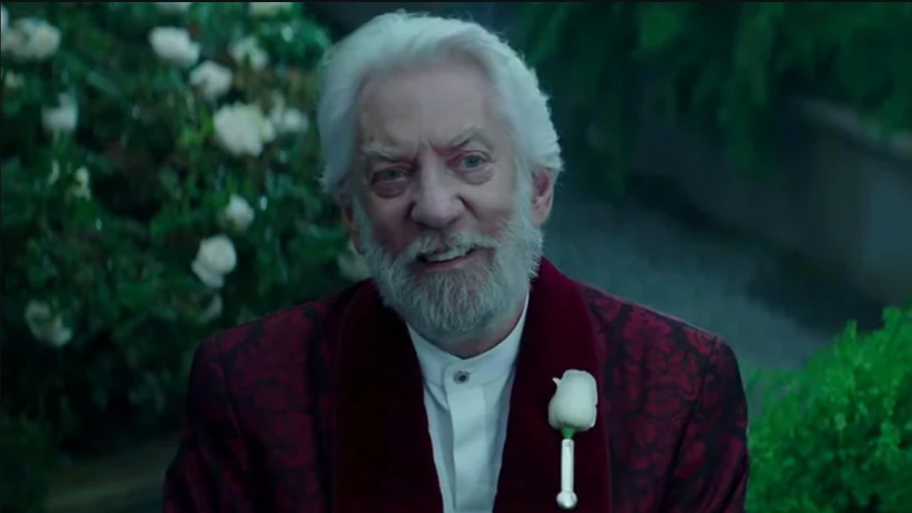 An older man with a white beard smiles, wearing a formal dark patterned jacket and white shirt with a rose boutonniere, in a garden setting